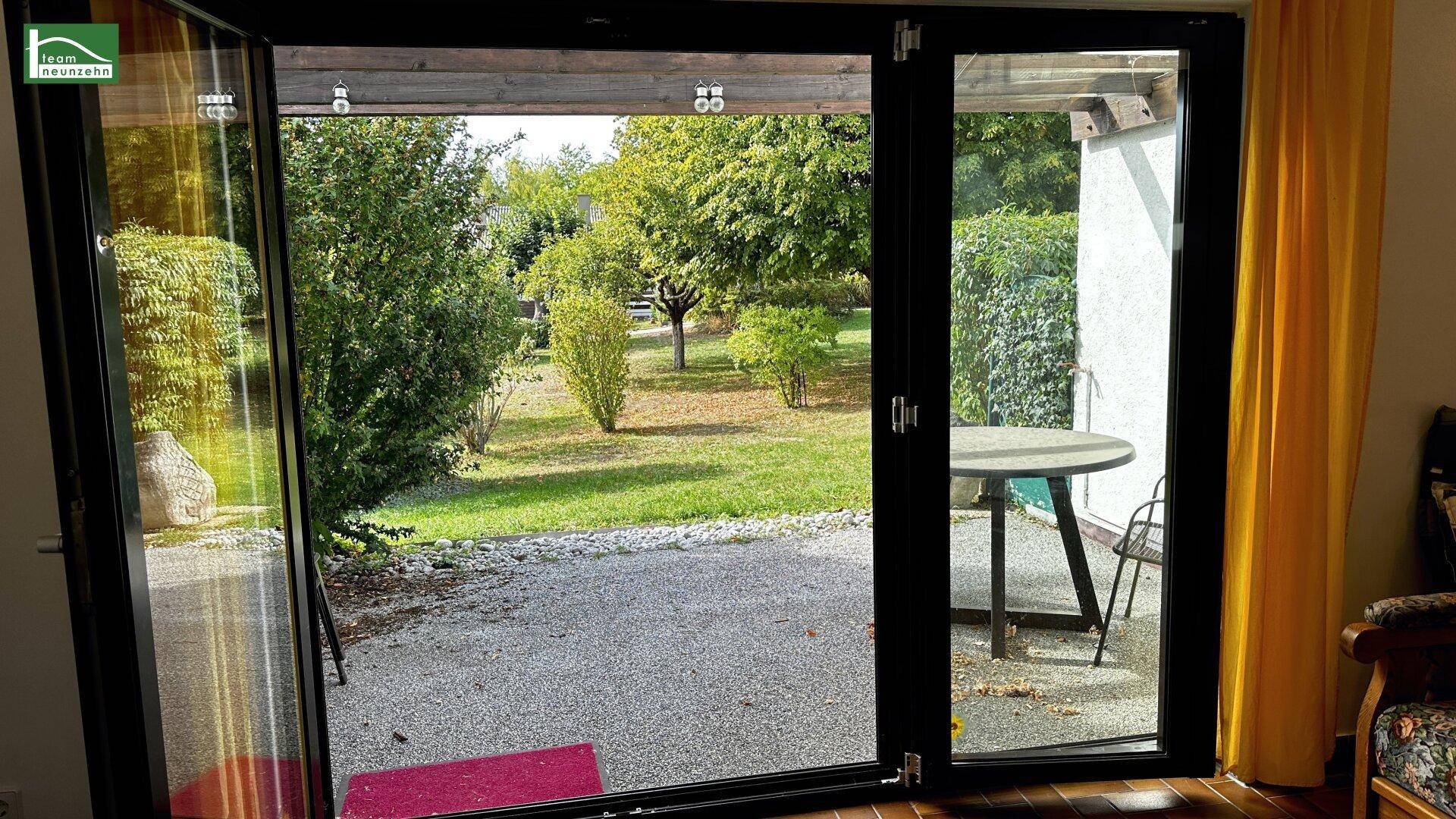Große Glastür bietet direkten Zugang zum Garten mit Bäumen und Sitzgelegenheit im Freien.