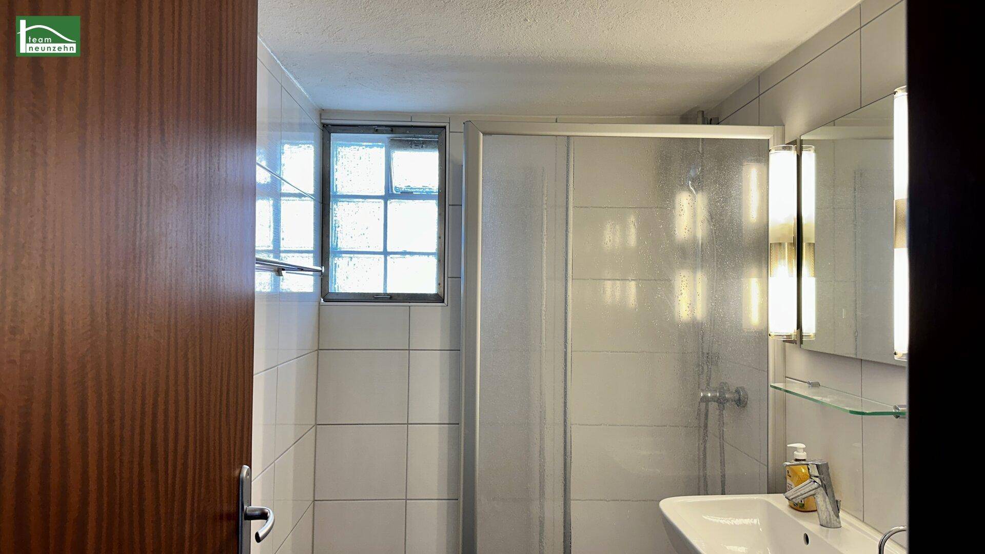 Badezimmer mit Duschkabine, Waschbecken und Fenster, hell gefliest.