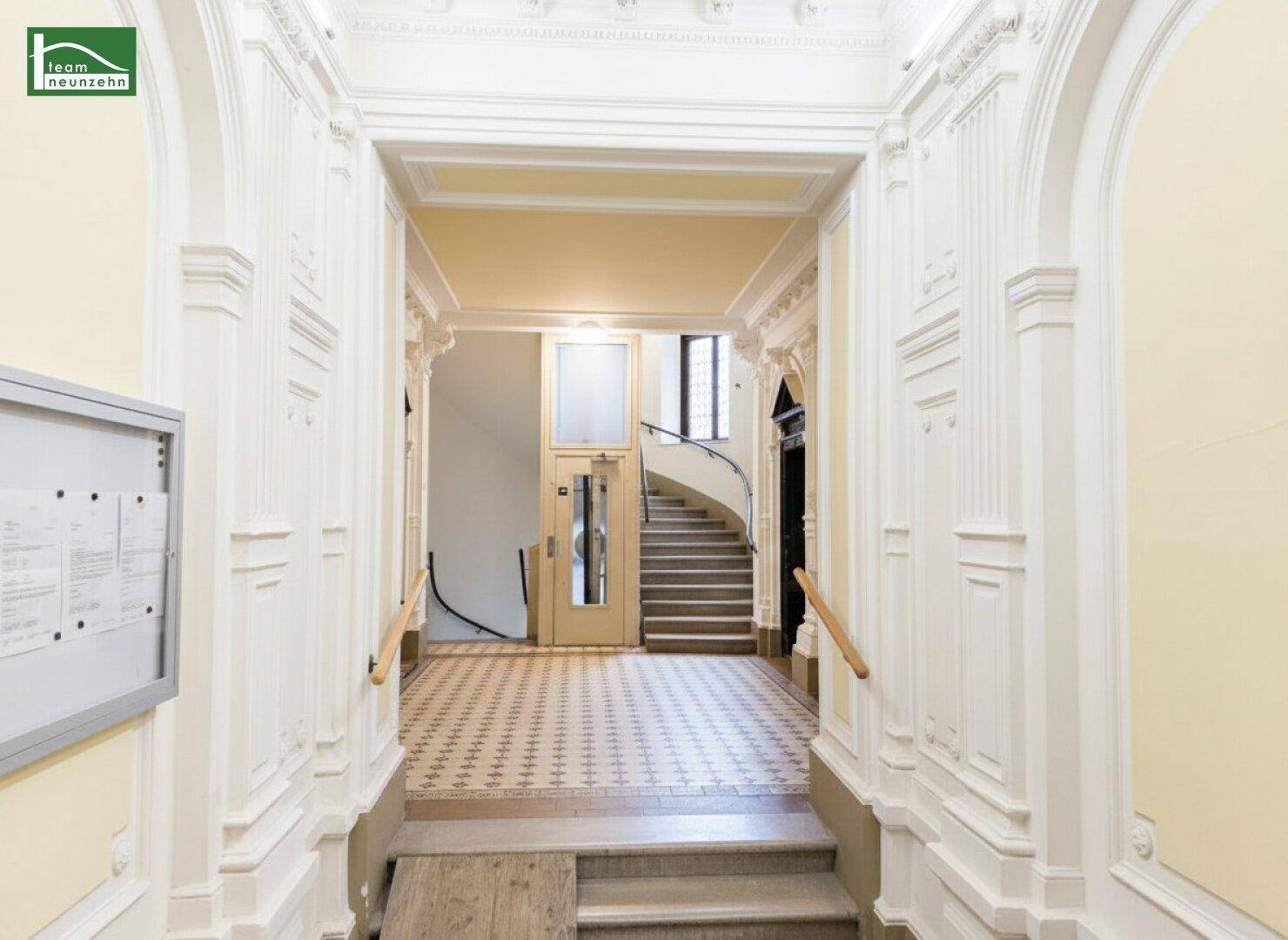Eleganter Flur mit historischem Aufzug, geschwungener Treppe und Stuckverzierungen.