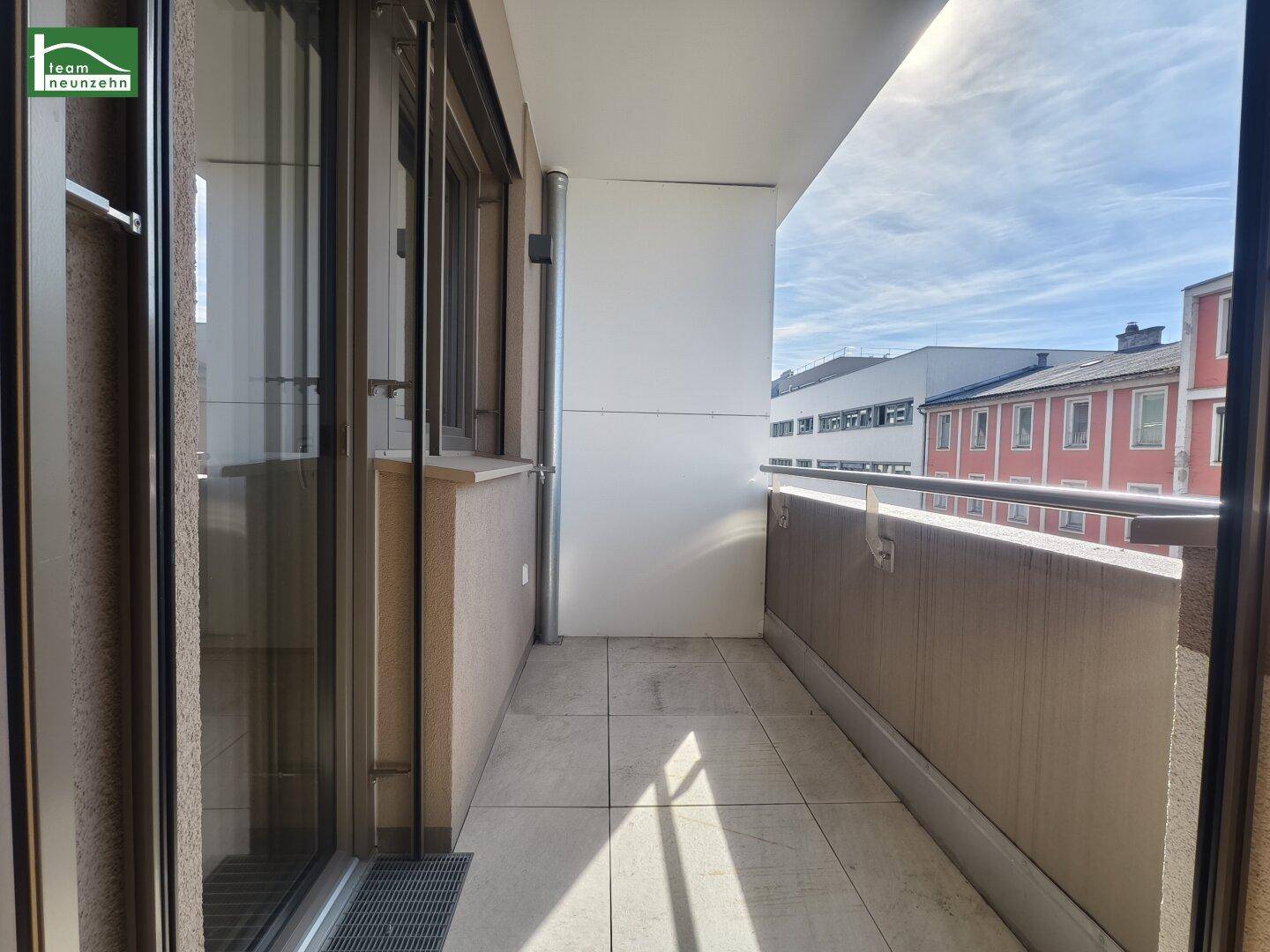 Schmaler Balkon mit Fliesenboden und Blick auf umliegende Gebäude unter blauem Himmel.