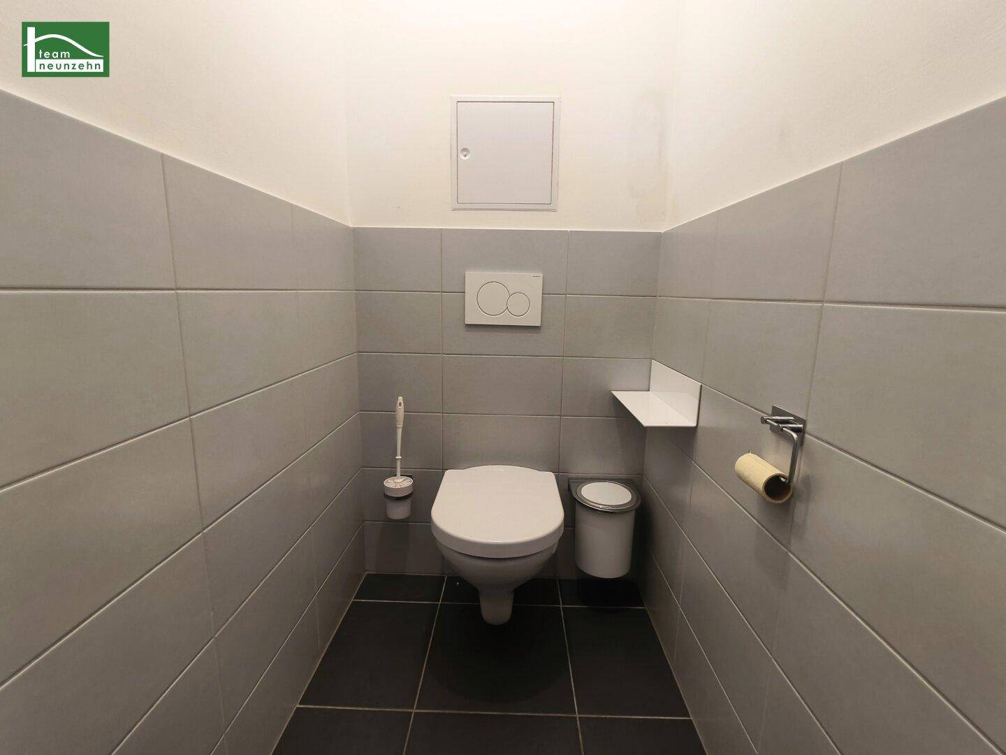 Separates WC mit grauen Fliesen, wandhängender Toilette und kleinem Waschbecken.