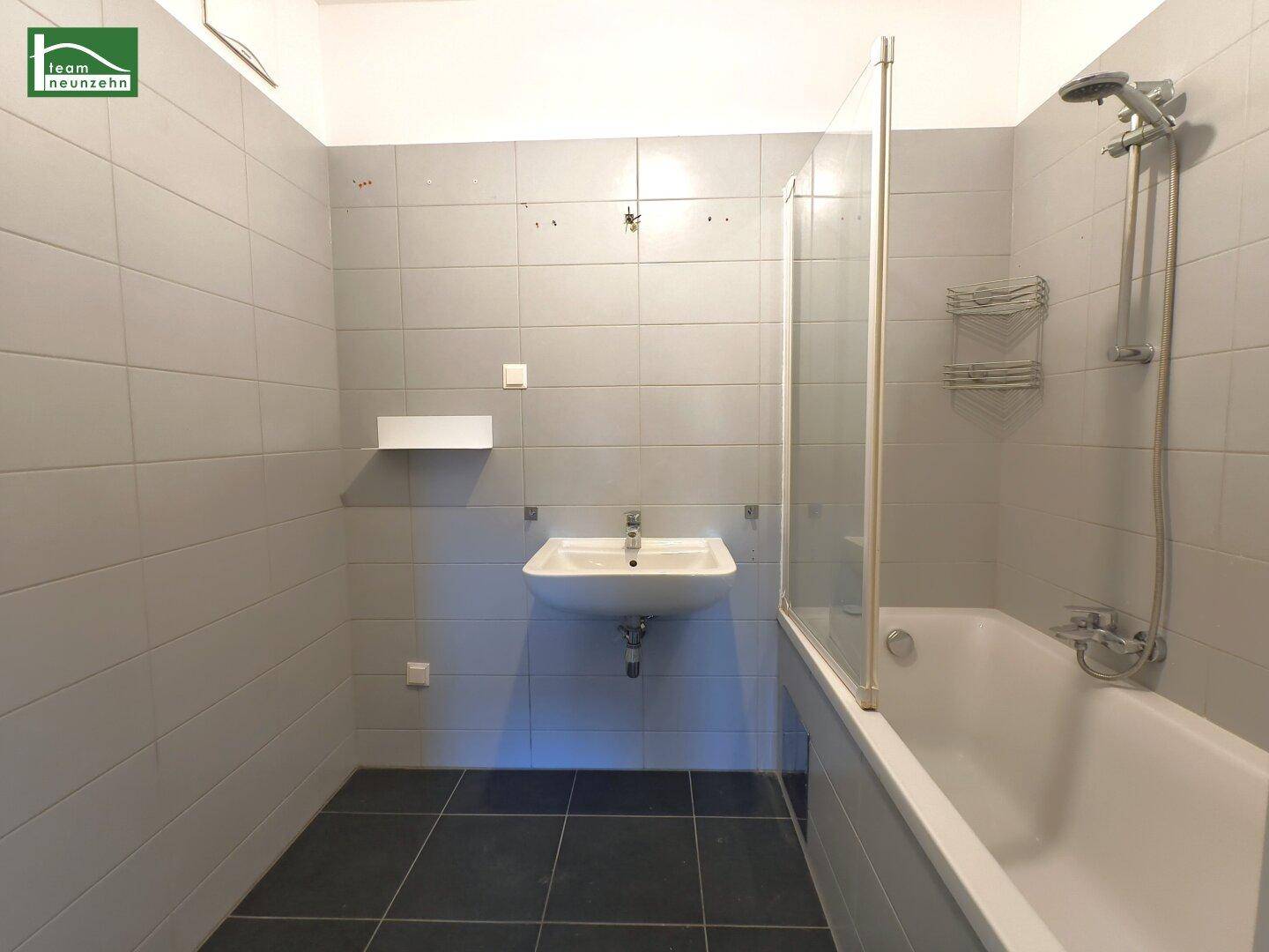 Modernes Badezimmer mit grauen Fliesen, Badewanne mit Duschwand und wandhängendem Waschbecken.