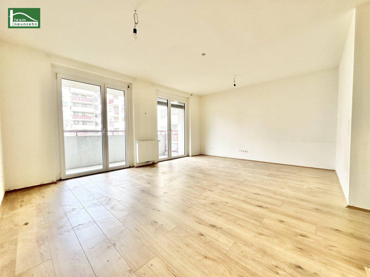 Helles, unmöbliertes Wohnzimmer mit Laminatboden und zwei großen Balkontüren, die viel Tageslicht hereinlassen.