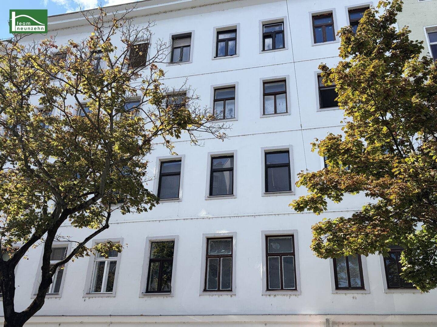 Weiße Fassade eines Mehrfamilienhauses mit vielen Fenstern und Bäumen im Vordergrund.