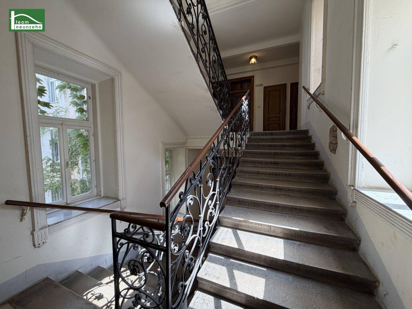 Historisches Treppenhaus mit schmiedeeisernem Geländer und großen Fenstern, die viel Licht hereinlassen.