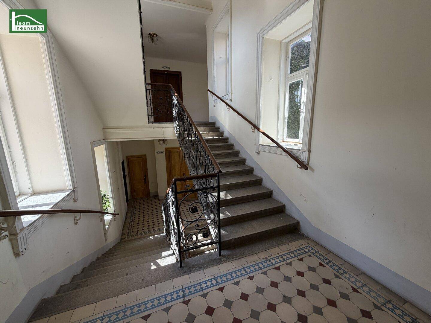 Historisches Treppenhaus mit schmiedeeisernem Geländer, großen Fenstern und gemustertem Fliesenboden.