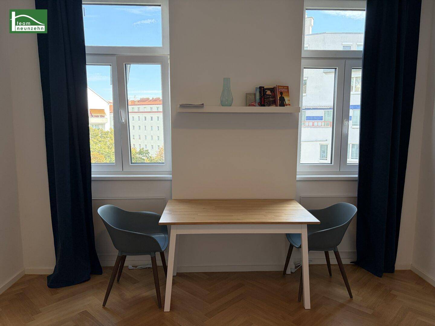 Helles Zimmer mit kleinem Esstisch und zwei Stühlen, umgeben von großen Fenstern mit dunklen Vorhängen.