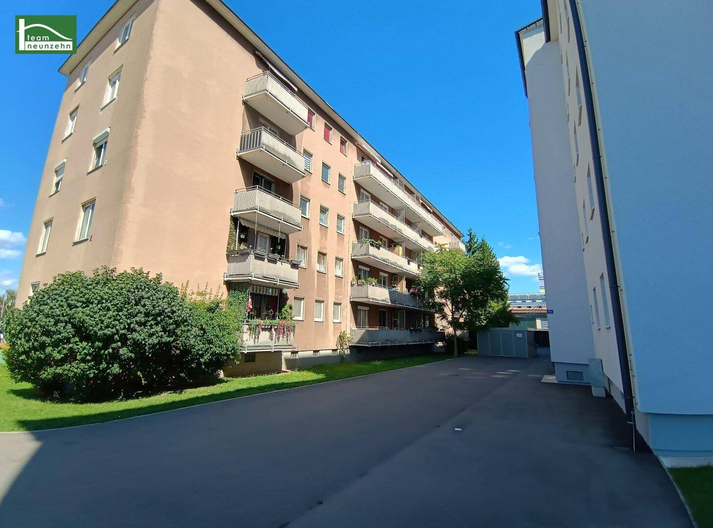 Mehrfamilienhaus mit Balkonen und grüner Umgebung unter blauem Himmel.
