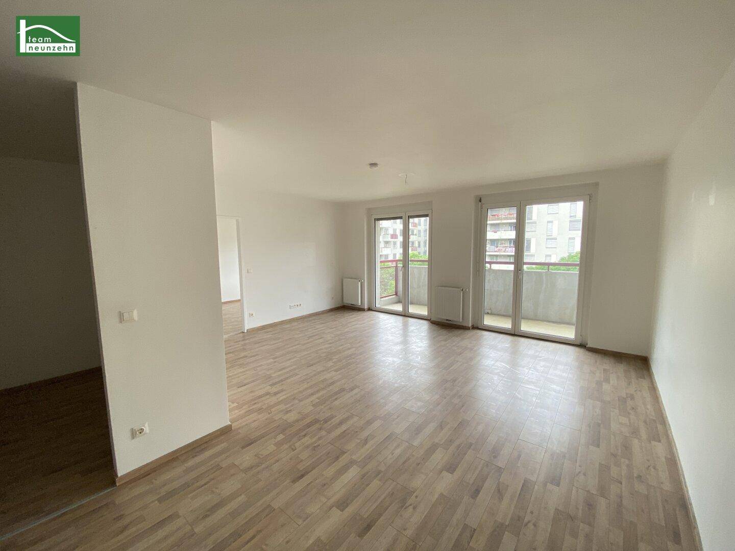 Geräumiger, leerer Wohn- und Essbereich mit hellem Holzboden und zwei Balkonzugängen.