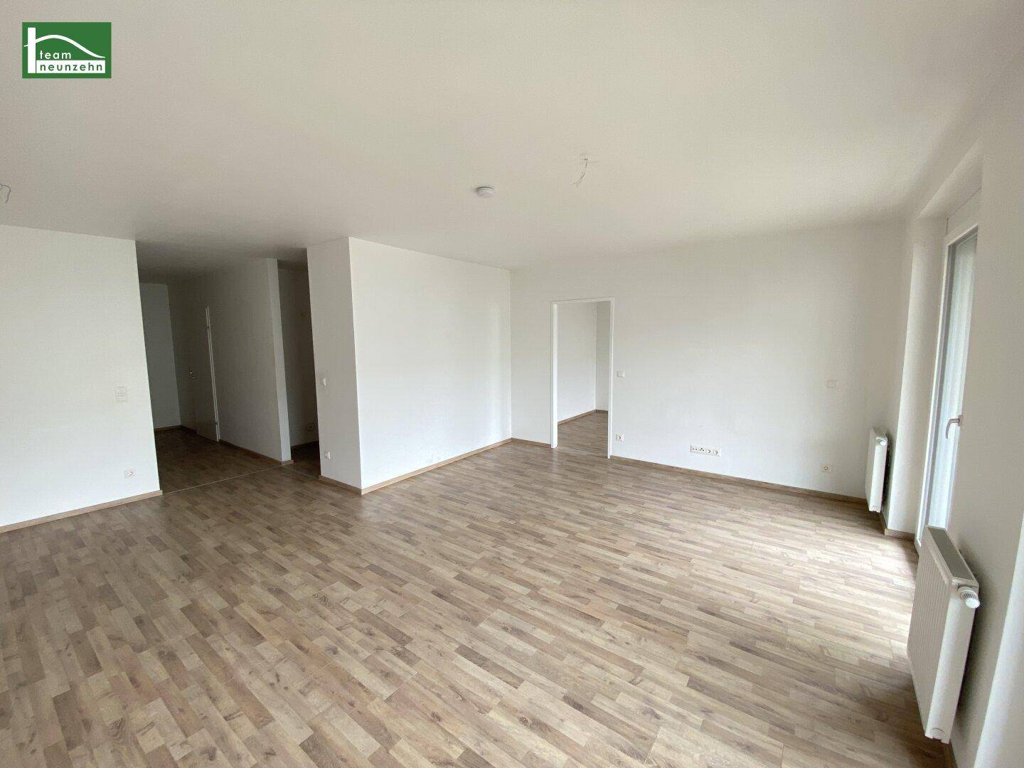 Heller, leerer Wohnbereich mit Holzboden und Zugang zu weiteren Zimmern.