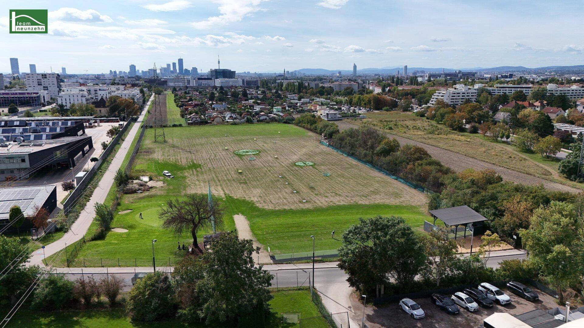 Weitläufige Luftaufnahme einer grünen Landschaft mit Golfplatz oder Park, umgeben von städtischer Bebauung in der Ferne.