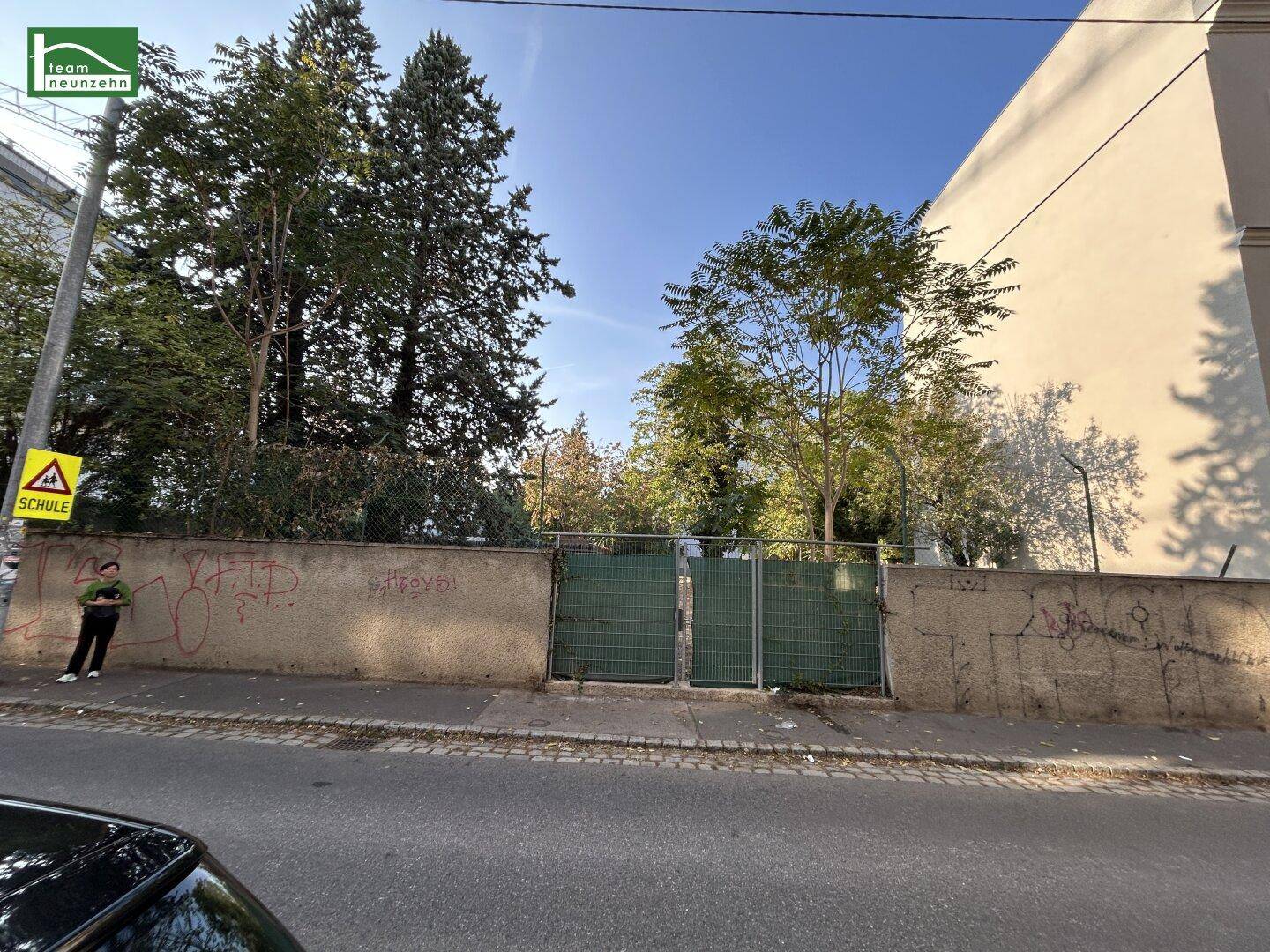 Straßenansicht mit einer Mauer, einem grünen Zaun und einem Schulschild am Straßenrand.