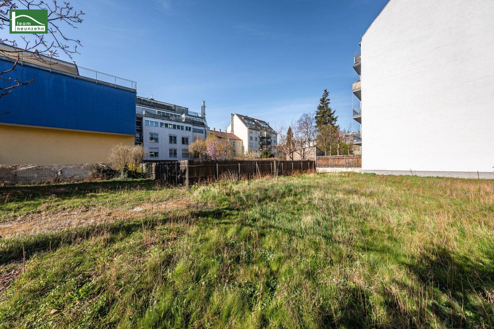 Grüne Wiese eines unbebauten Grundstücks mit umliegenden Gebäuden unter blauem Himmel.