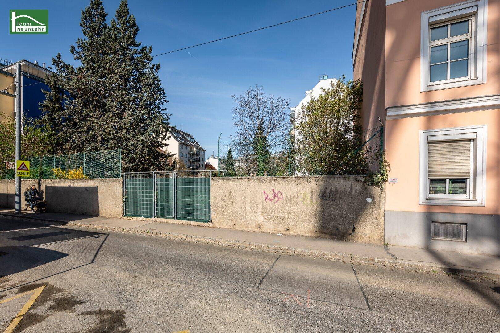 Straßenansicht eines Grundstücks mit Mauer, grünem Tor und einem Schulschild.