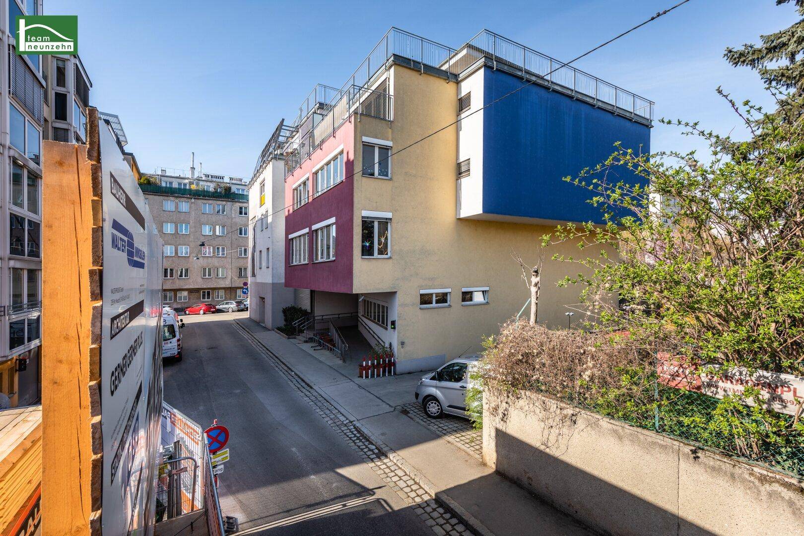 Gebäudefassade mit blauen und roten Akzenten, angrenzender Straße und weiteren Häusern.
