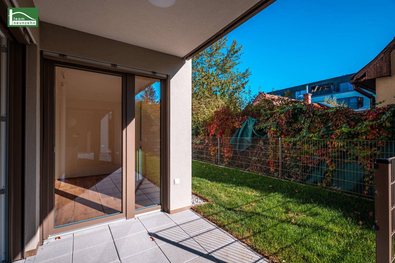Sonniger Terrassenbereich mit Steinplatten und Zugang zu einem kleinen Garten, umgeben von grünen Hecken.