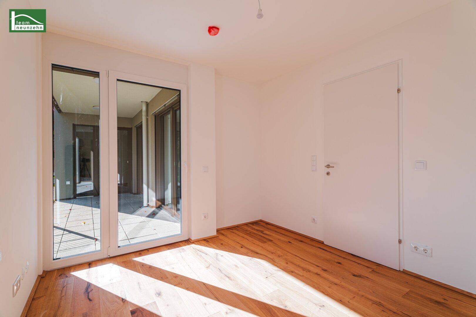 Heller Raum mit Holzboden und großen Glastüren, die viel Sonnenlicht hereinlassen und Zugang nach draußen bieten.