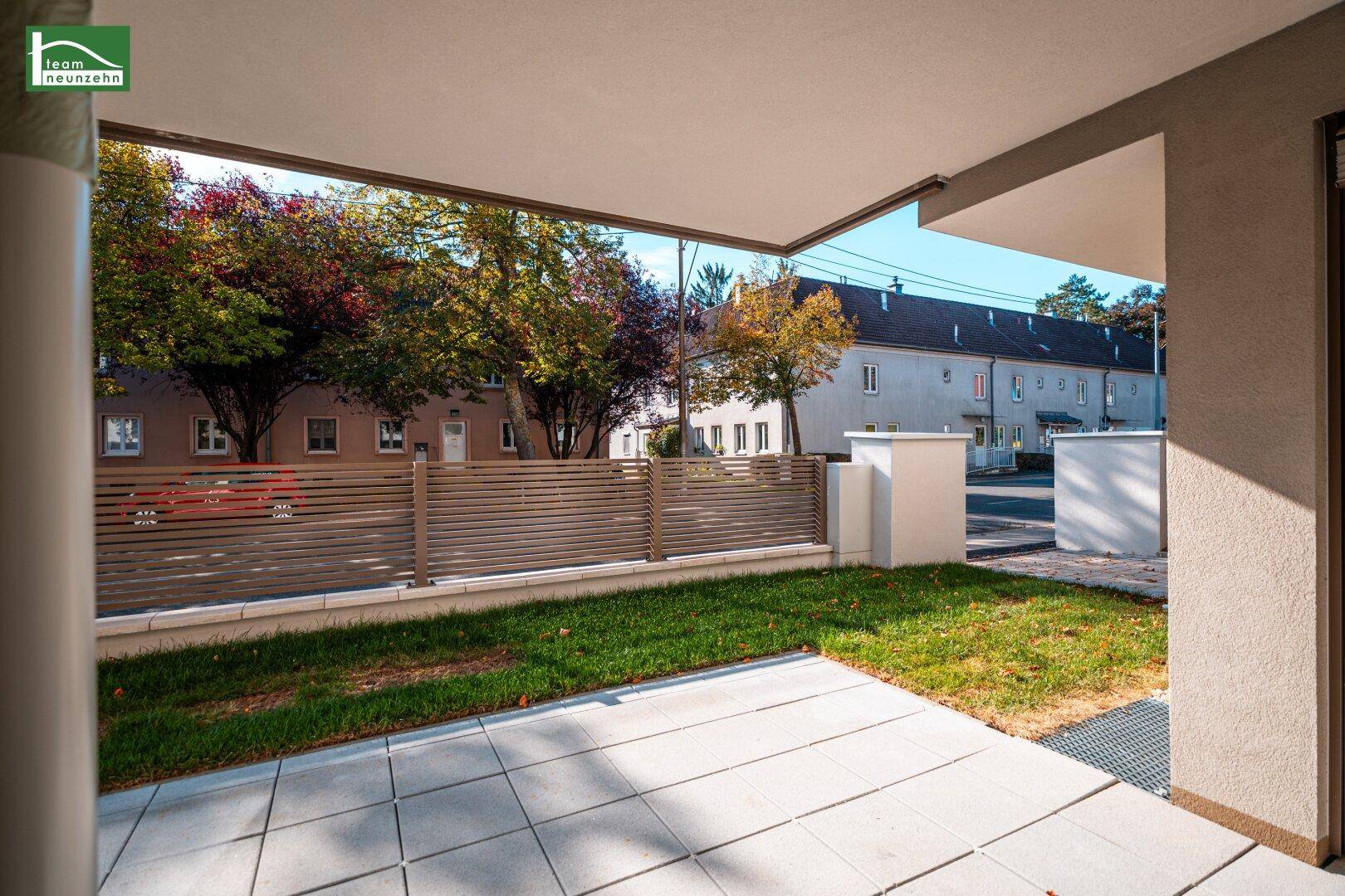 Überdachte Terrasse mit Steinplatten und kleinem Rasenbereich, umgeben von einem Zaun.