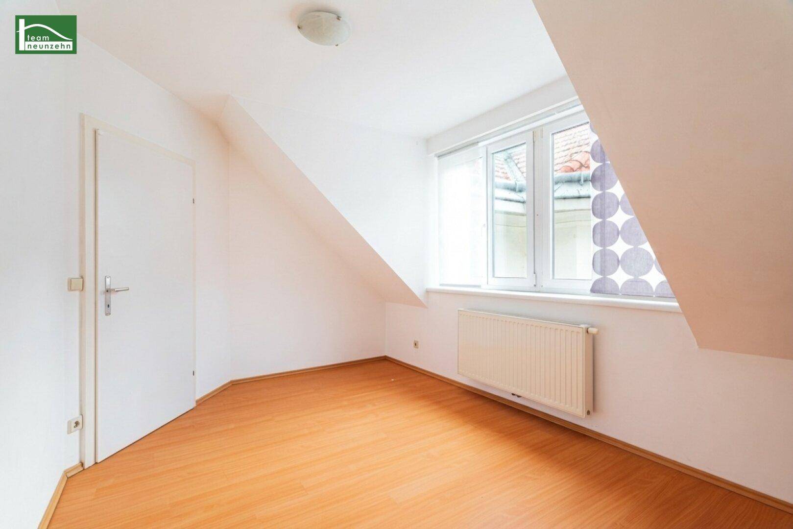 Helles Zimmer im Dachgeschoss mit Holzboden, Heizkörper und Fenster mit Blick nach draußen.