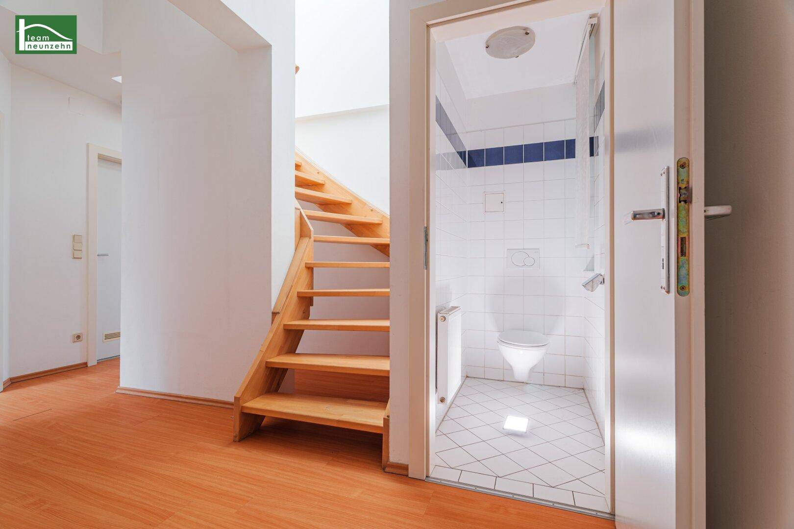 Flur mit Holztreppe und Zugang zu einem gefliesten WC, das durch eine Tür sichtbar ist.