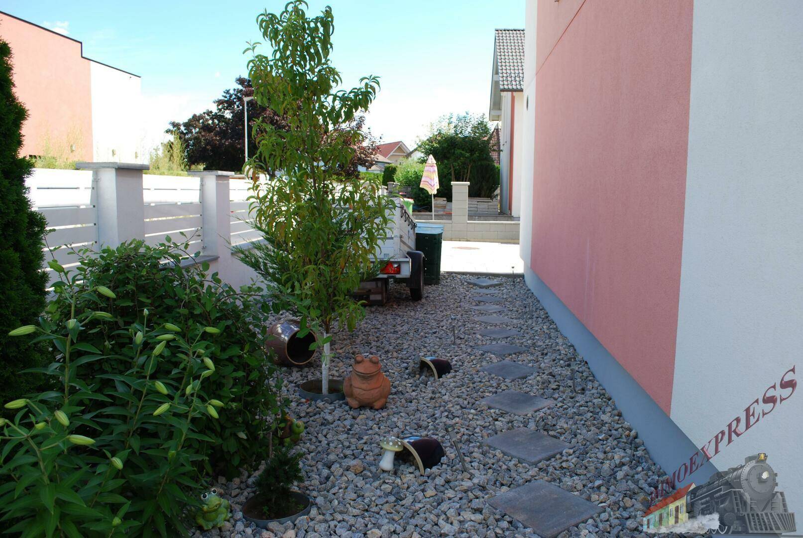 Seitlicher Kiesweg entlang des Hauses mit dekorativen Pflanzen und Elementen.