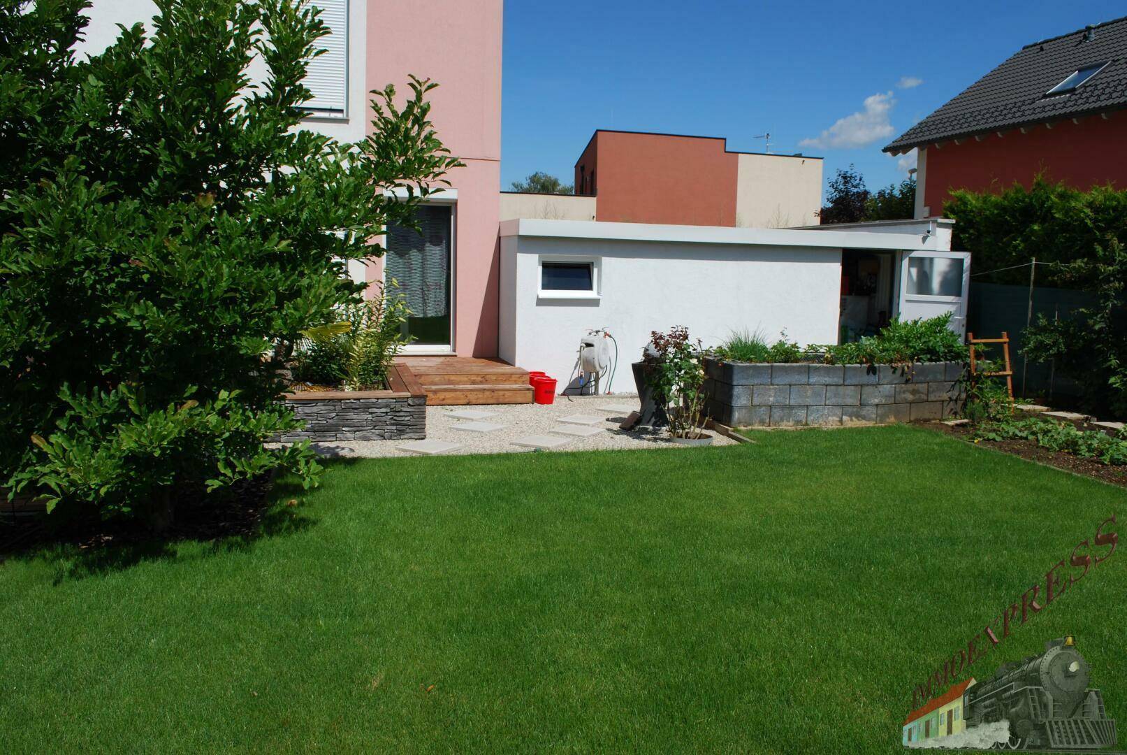 Rückseite des Hauses mit Rasenfläche, kleiner Terrasse und bepflanzten Hochbeeten.