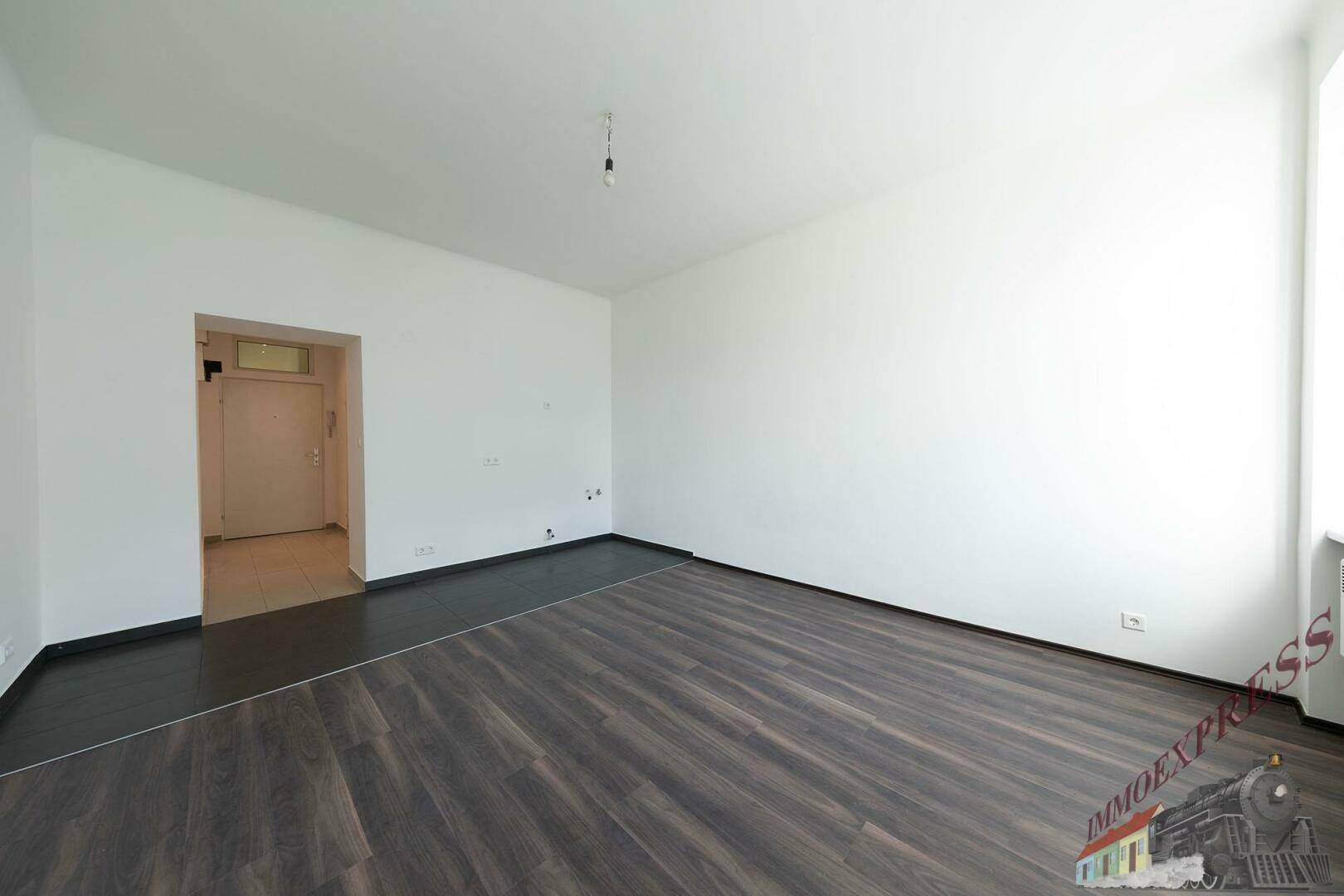 Geräumiges, leeres Zimmer mit dunklem Laminatboden und Zugang zu einem weiteren Bereich.