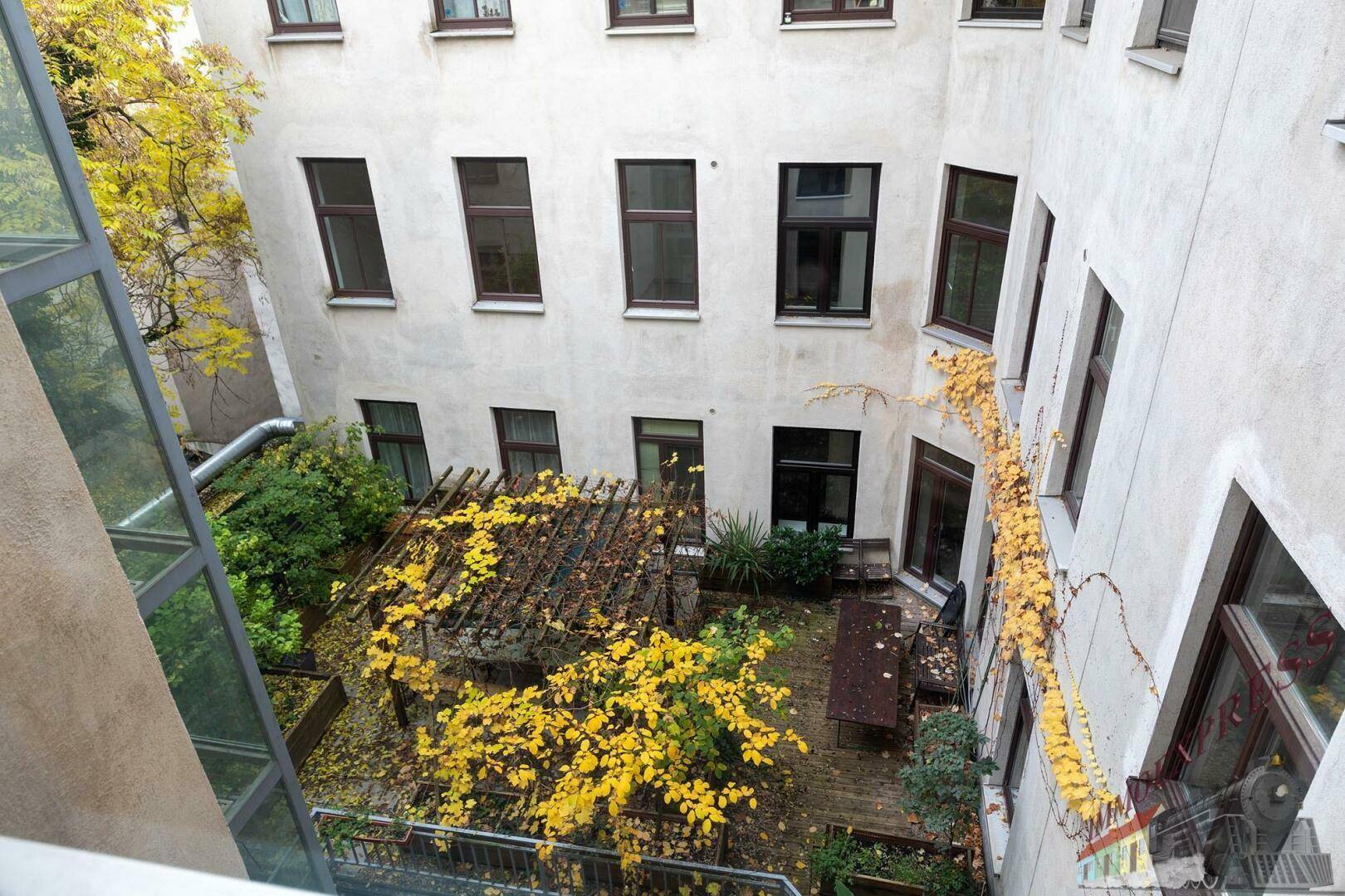 Blick von oben auf einen grünen Innenhof mit Pergola und Sitzgelegenheiten im Herbstlaub.