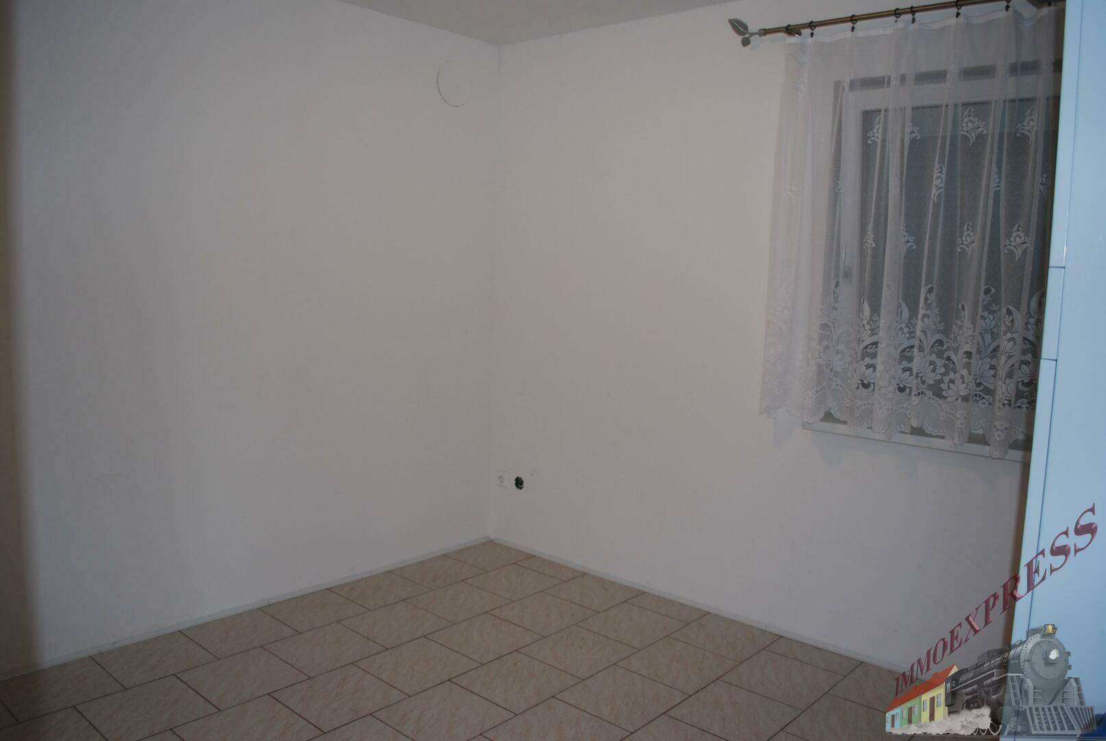 Leeres Zimmer mit Fliesenboden und einem Fenster mit Gardinen.