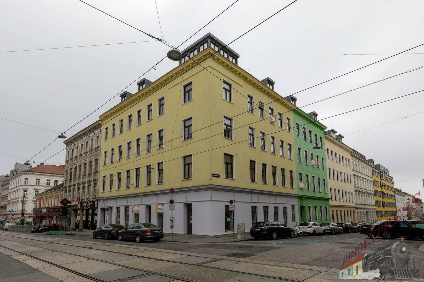 Eckgebäude mit gelber Fassade an einer belebten Straße mit Straßenbahnschienen.