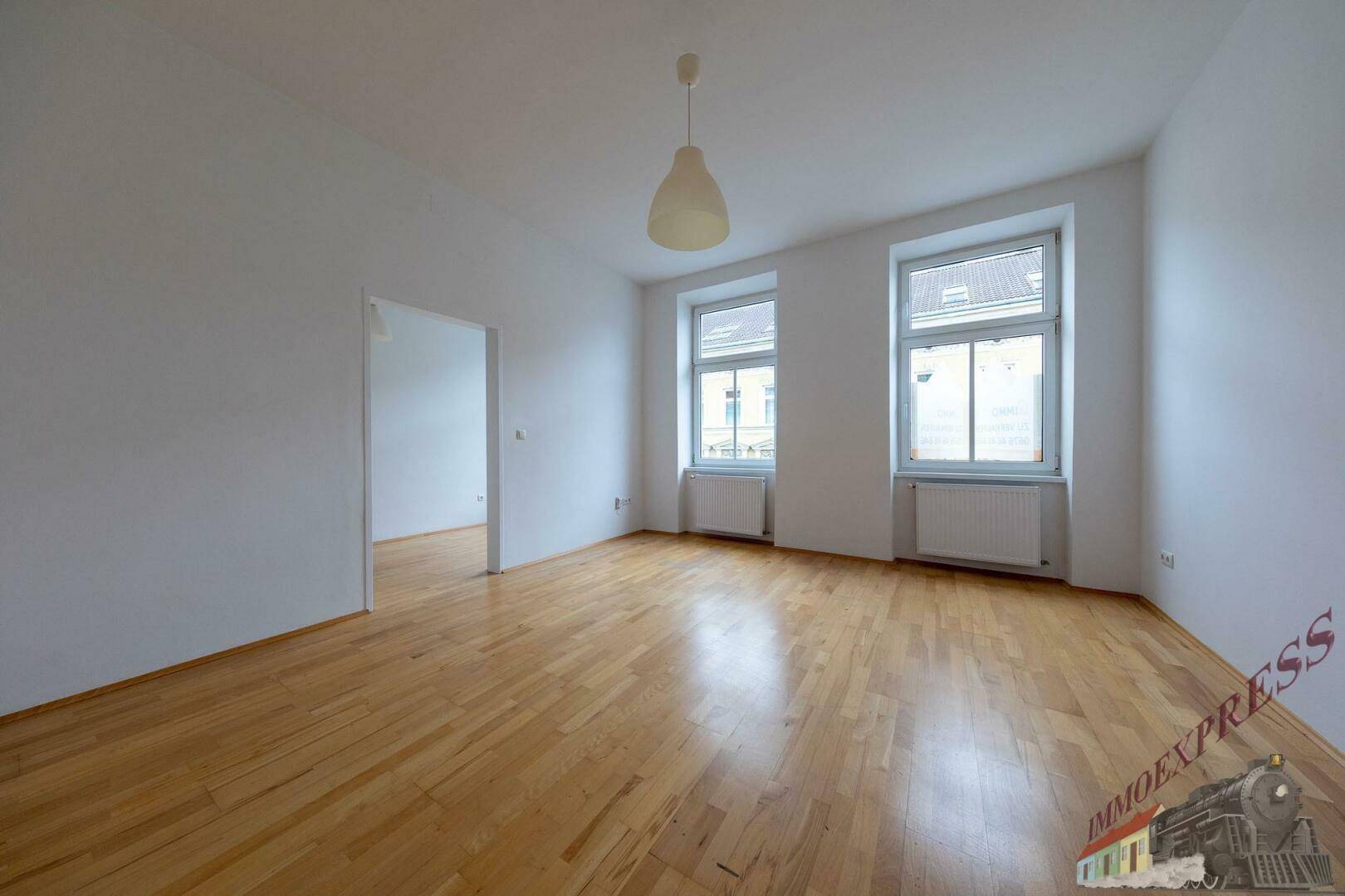 Geräumiges Wohnzimmer mit hellem Parkettboden und zwei großen Fenstern.