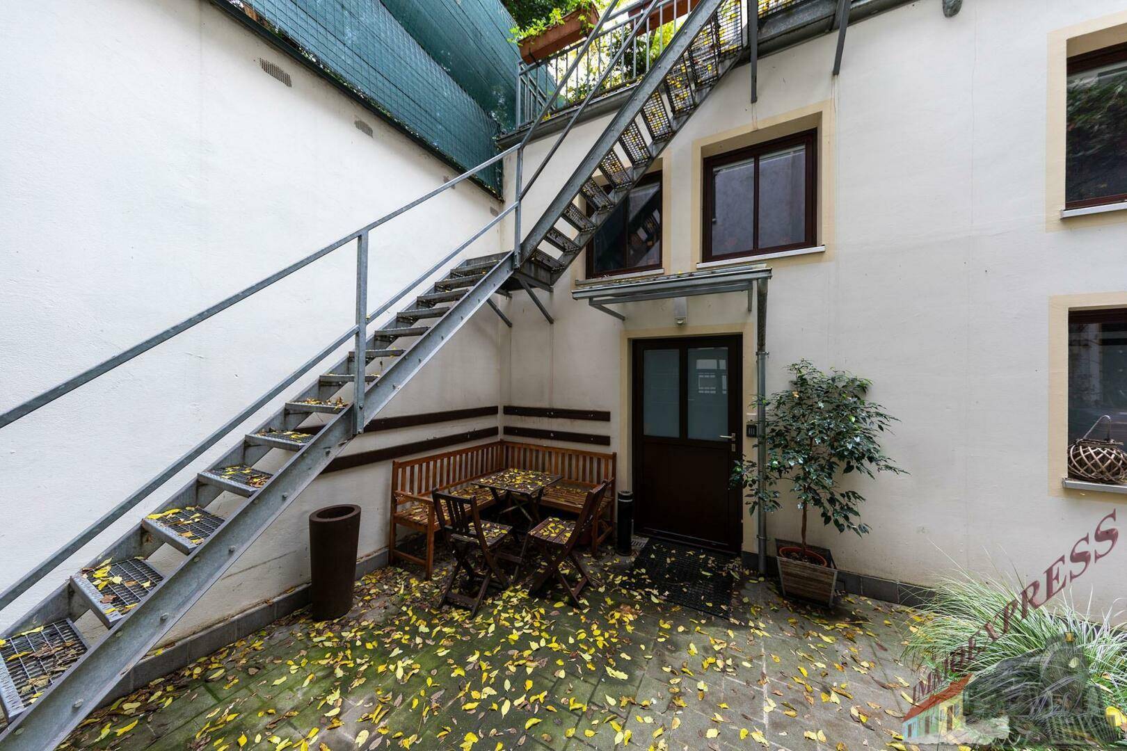 Kleiner Innenhof mit Sitzgelegenheiten, Außentreppe und herbstlichem Laub auf dem Boden.