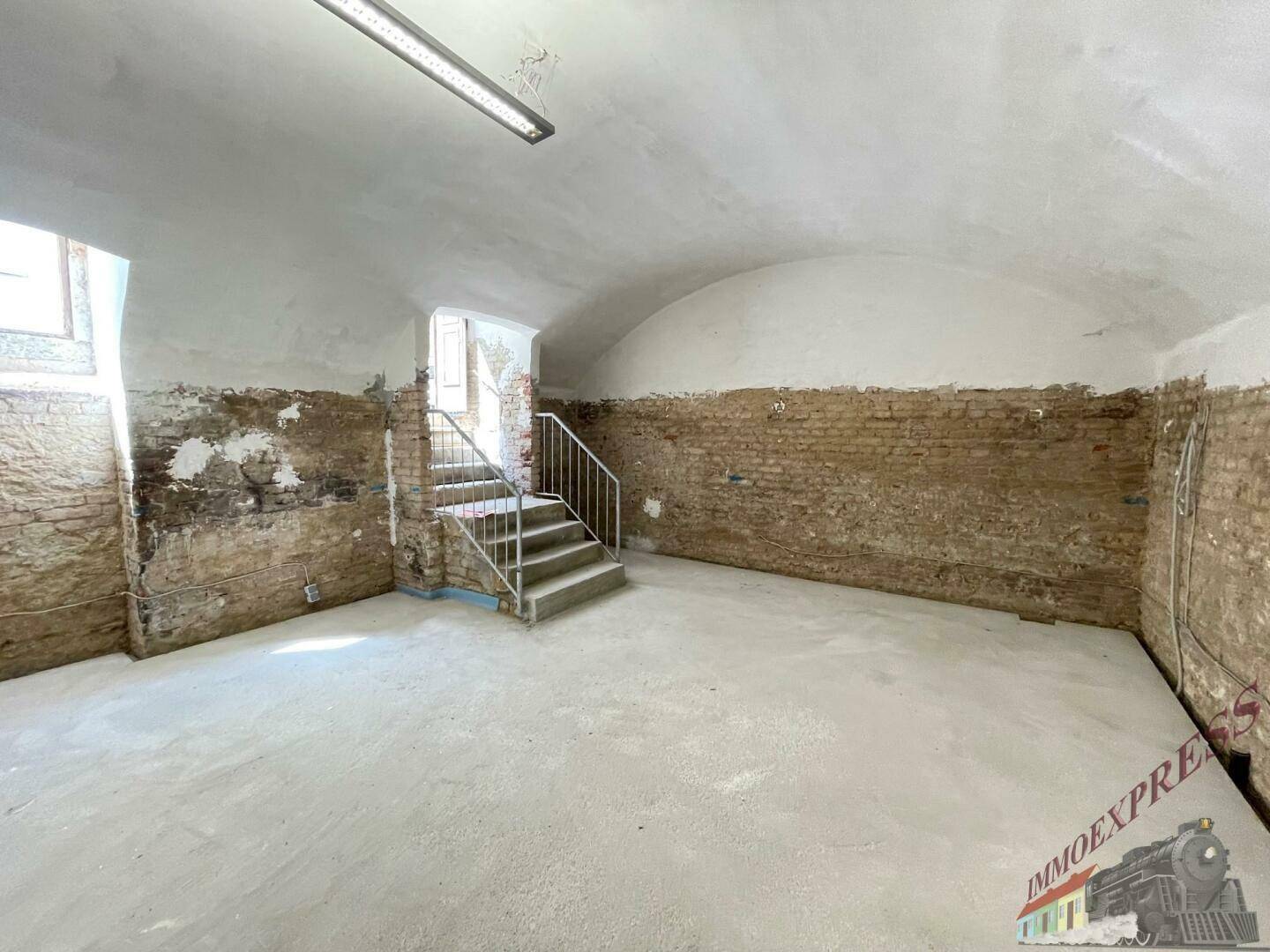 Innenansicht eines großen Raumes mit Gewölbedecke, unverputzten Ziegelwänden und einer Treppe.