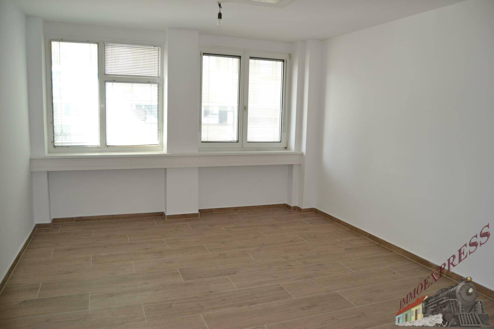 Geräumiger Büroraum mit großen Fenstern und hellem Laminatboden, bietet viel Tageslicht.