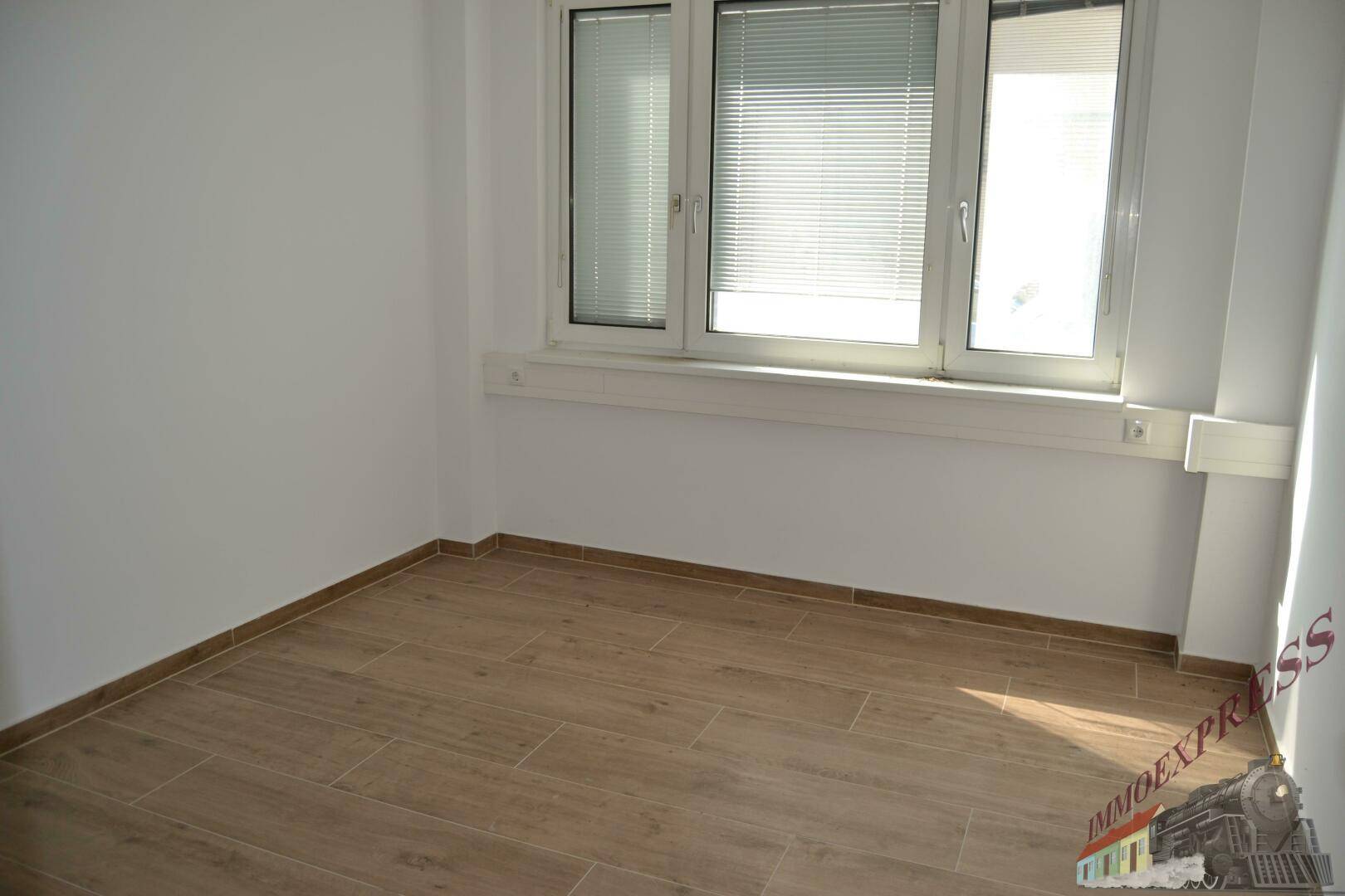 Heller Büroraum mit einem Fenster, Jalousien und einem modernen Laminatboden in Holzoptik.