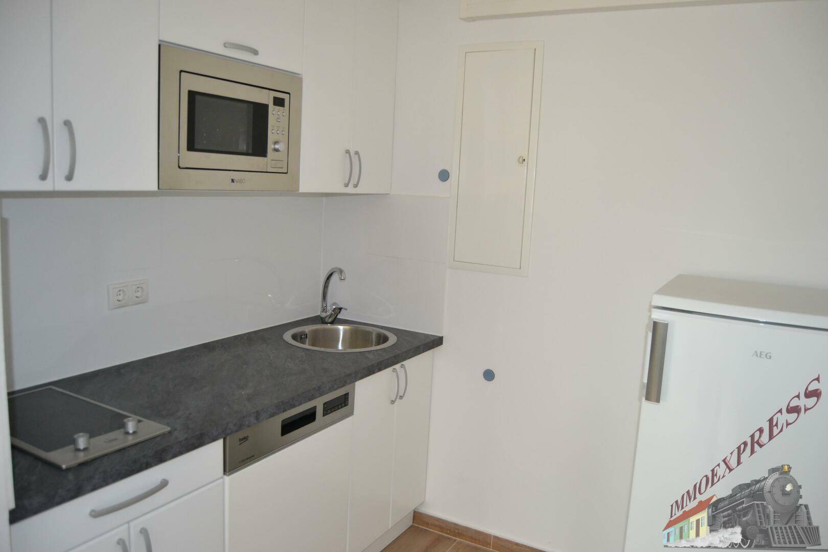 Kompakte Küchenzeile mit weißen Schränken, Spüle, Kochfeld, Mikrowelle und Kühlschrank.