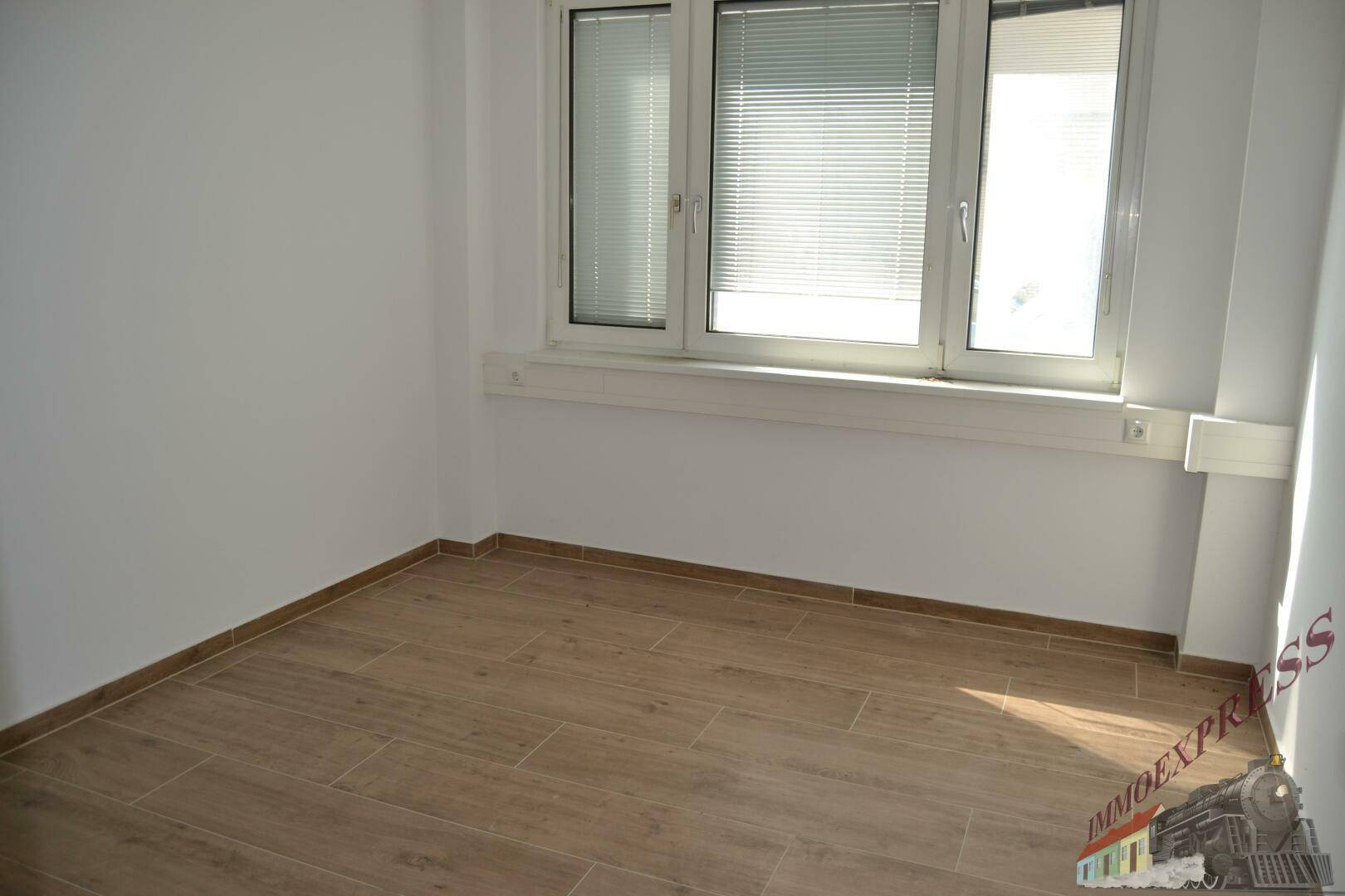 Heller Raum mit einem Fenster und Jalousien, ausgestattet mit einem modernen Fliesenboden in Holzoptik.