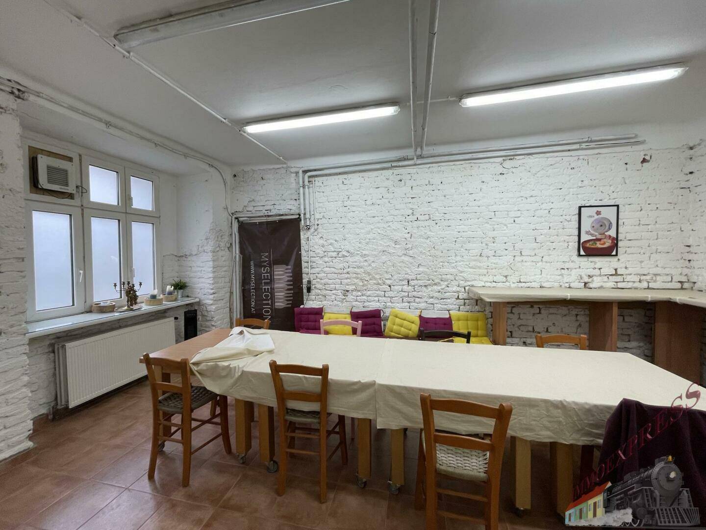 Großer Mehrzweckraum mit langen Tischen, Stühlen, weiß gestrichenen Ziegelwänden und Fenstern.