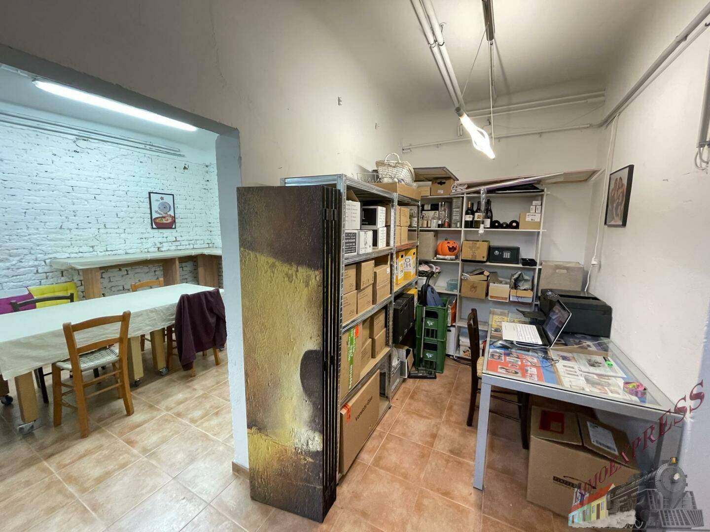 Kombinierter Büro- und Lagerbereich mit Regalen, Schreibtisch und Blick in den Mehrzweckraum.