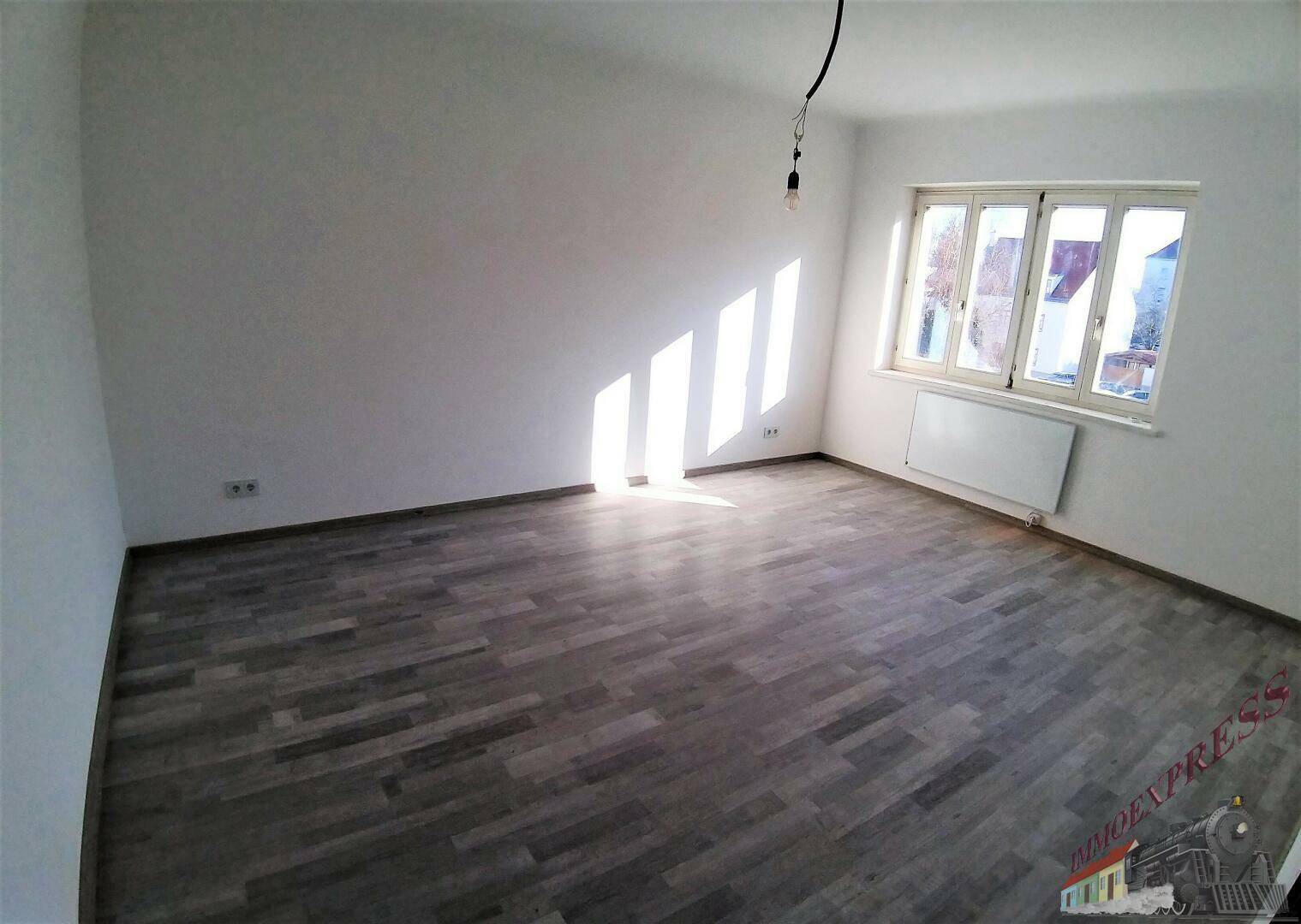 Leerer Raum mit dunklem Laminatboden und viel natürlichem Licht durch ein großes Fenster.