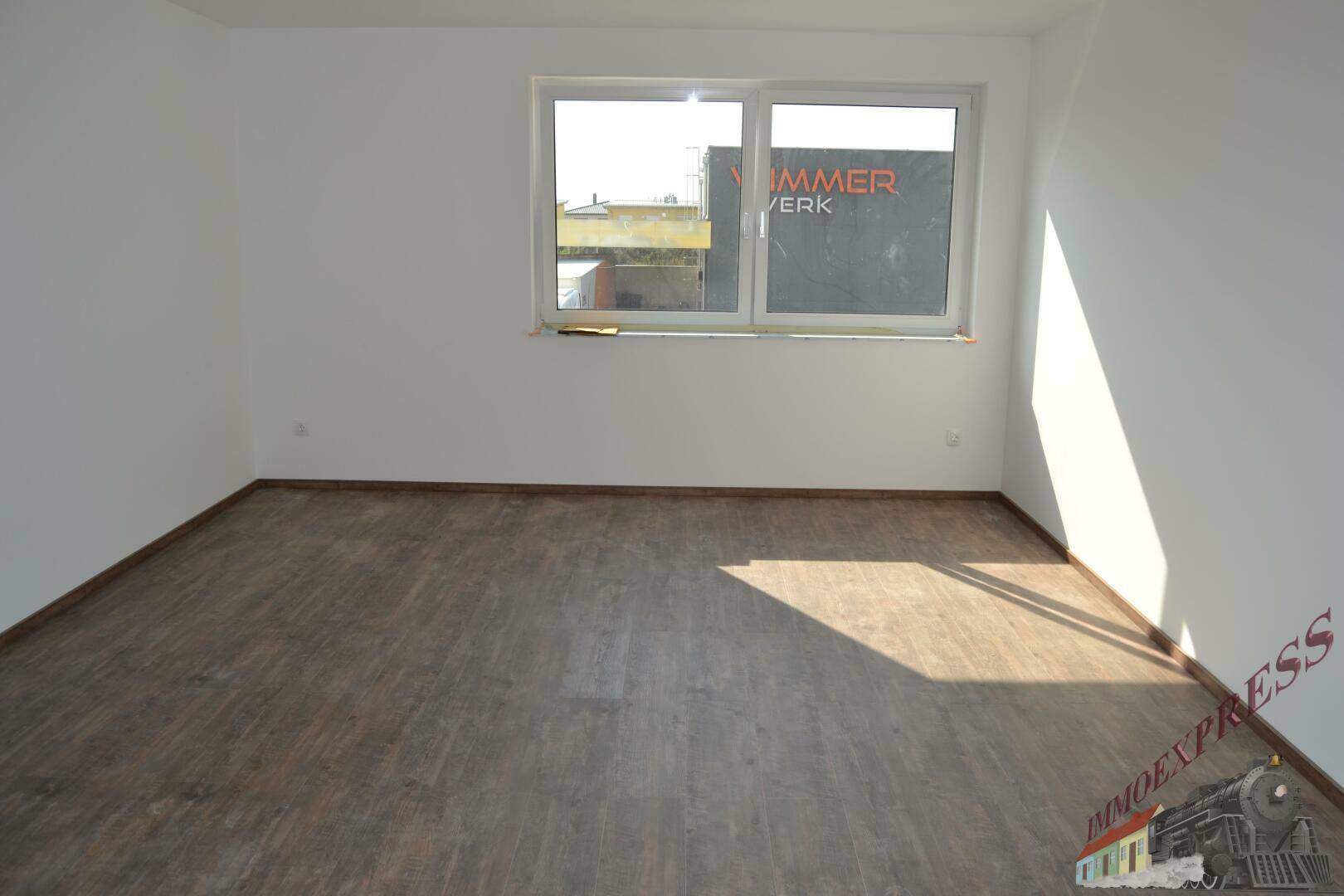 Leerer Raum mit Laminatboden und einem Fenster, das einen Blick auf ein Industriegebiet bietet.