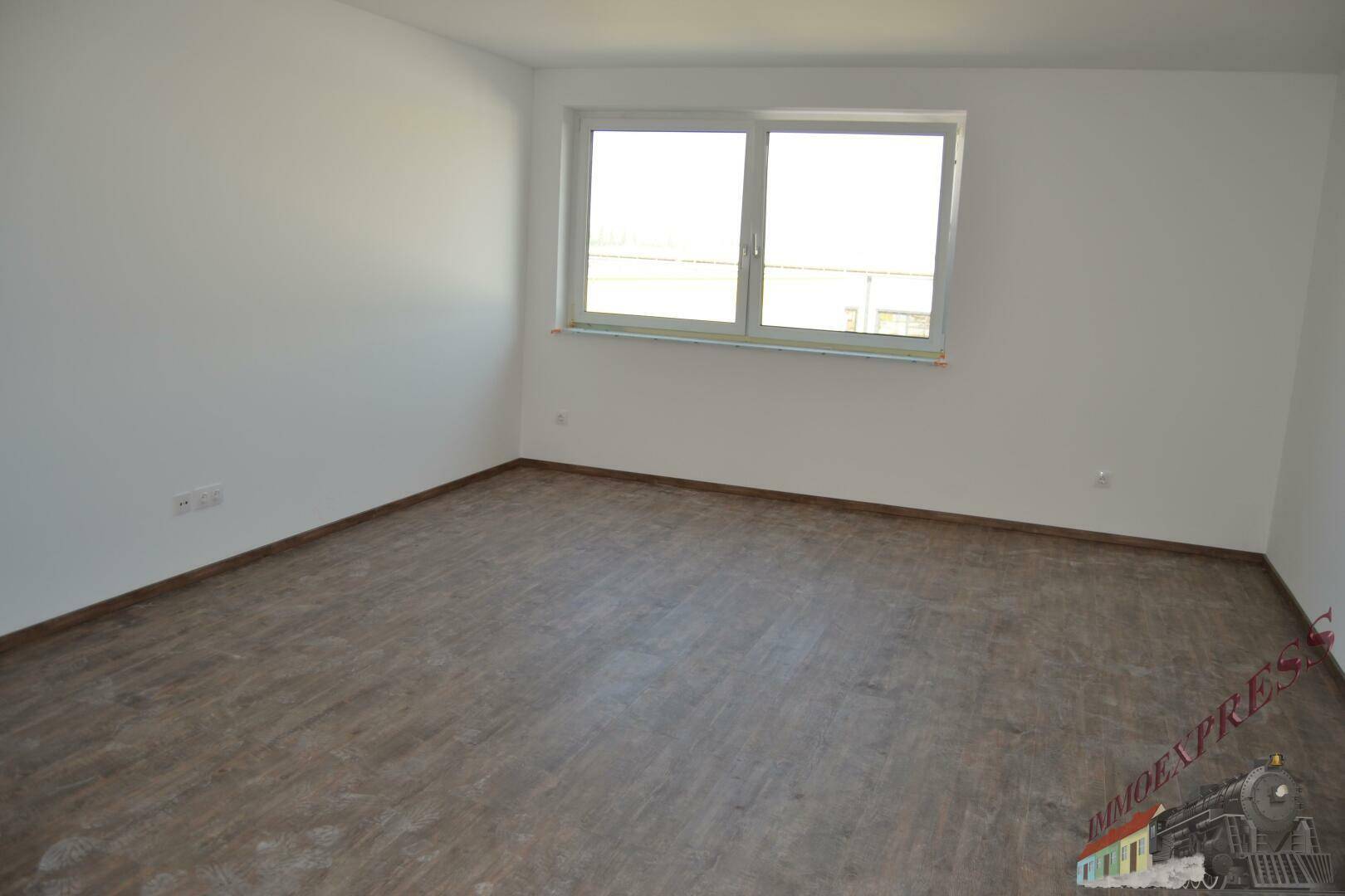 Heller, leerer Raum mit Laminatboden und einem großen Fenster, das viel Tageslicht hereinlässt.