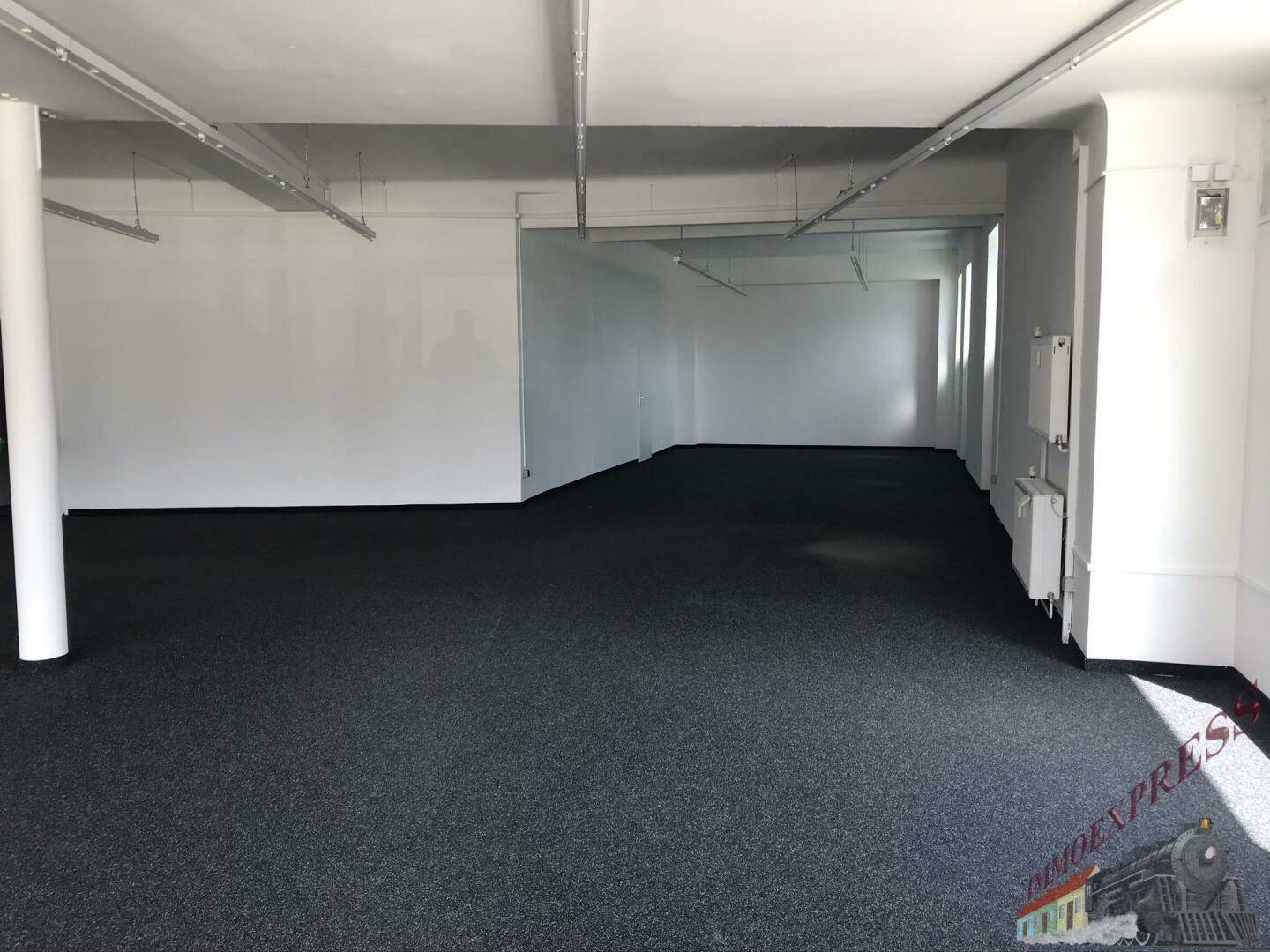 Geräumiger, leerstehender Verkaufsraum mit dunklem Teppichboden, weißen Wänden und Deckenbeleuchtung, bereit zur Nutzung.