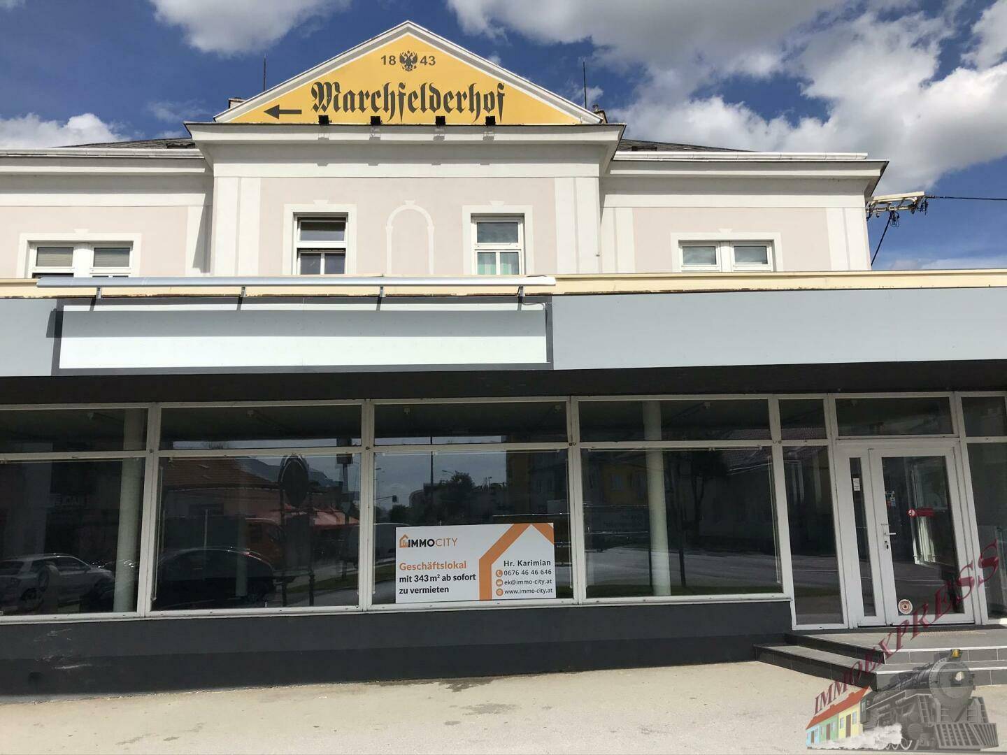 Ansicht der Geschäftsfront mit großen Schaufenstern und dem historischen "Marchfelderhof"-Schild darüber, unter blauem Himmel.