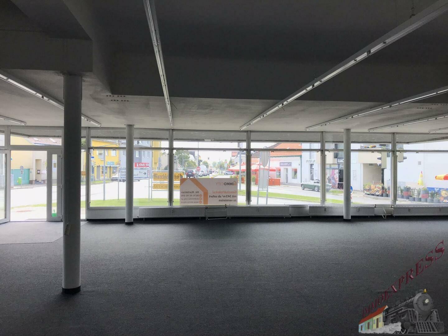 Geräumiger, leerer Geschäftsraum mit Teppichboden und großen Fenstern zur Straße, ausgestattet mit Deckenbeleuchtung.