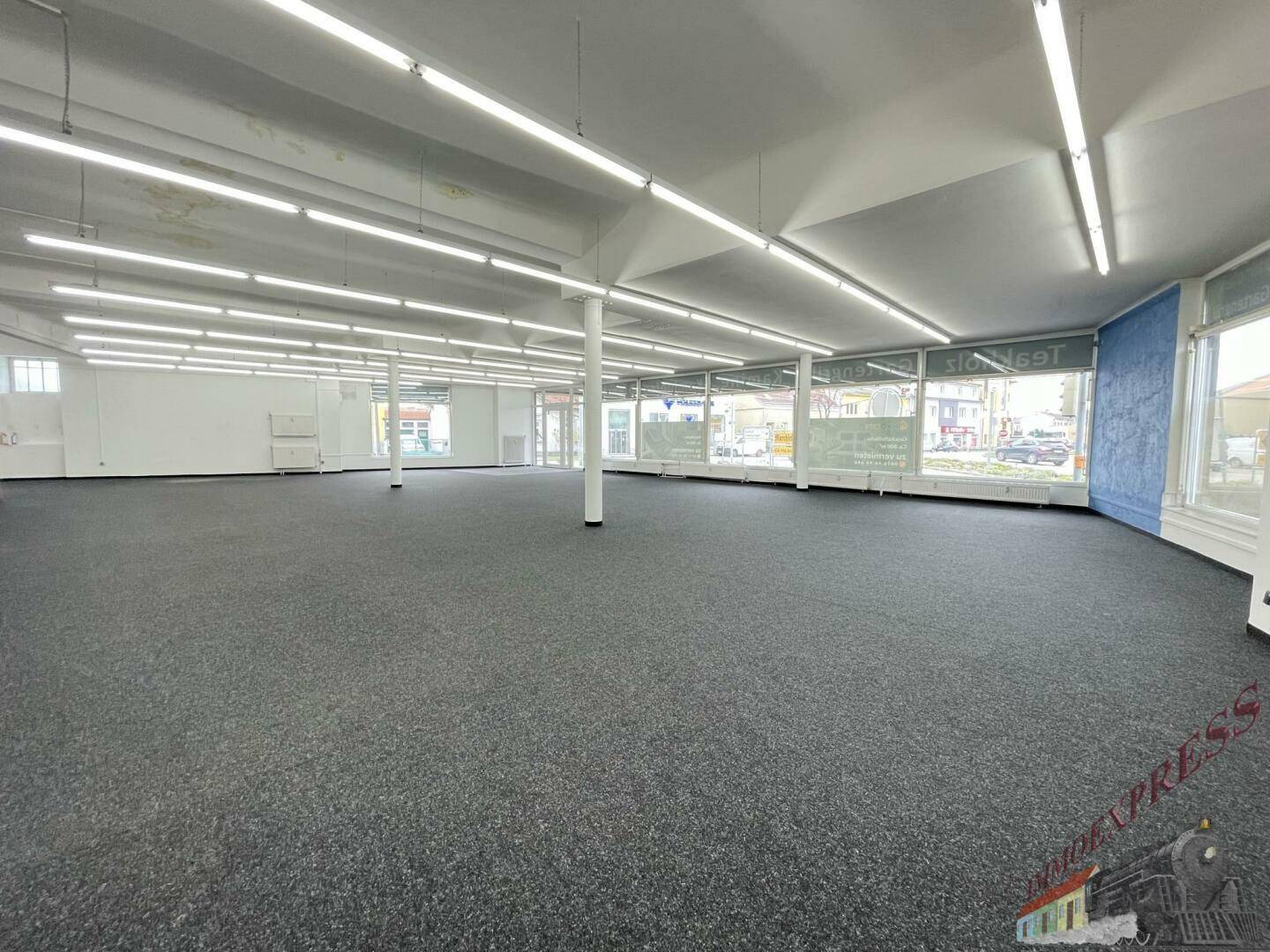 Weitläufiger Verkaufsraum mit dunklem Teppichboden, Säulen und durchgehenden Fensterfronten.