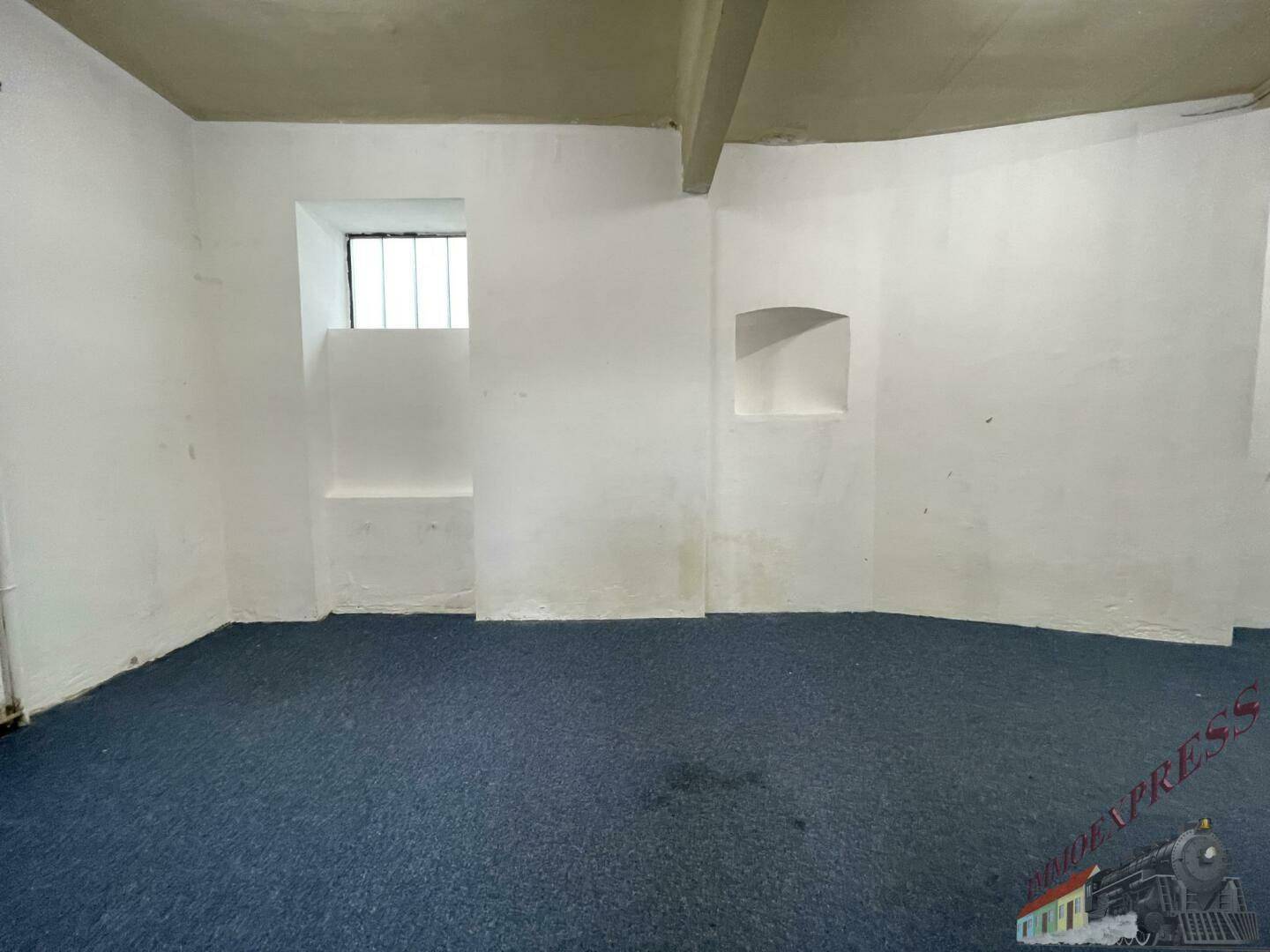 Einfacher Lagerraum mit blauem Teppichboden, kleinen vergitterten Fenstern und Nischen in der Wand.