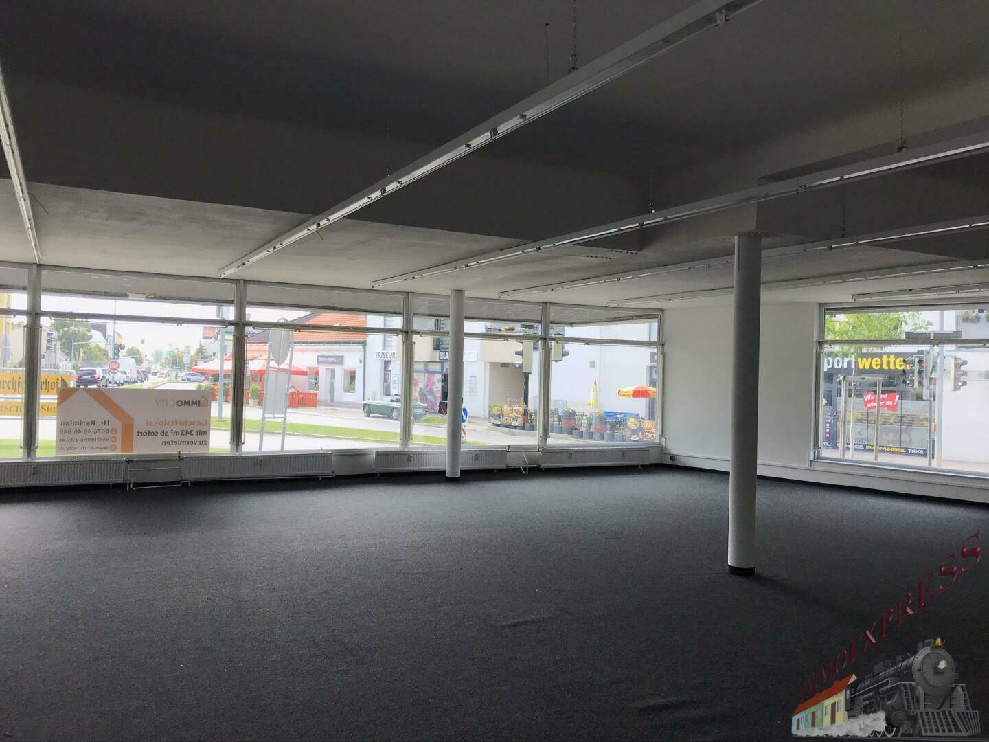 Weitläufiger Ladenbereich mit Teppichboden, Säulen und großen Fenstern zur Straße hin.