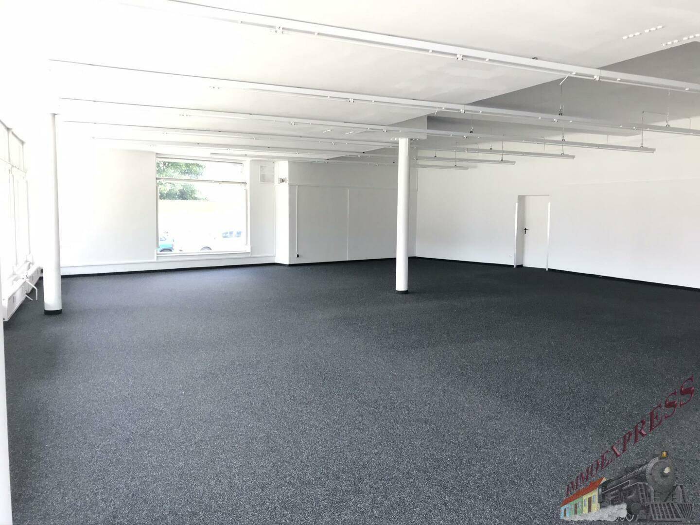 Heller, leerer Geschäftsraum mit Teppichboden, weißen Wänden und Deckenbeleuchtung.