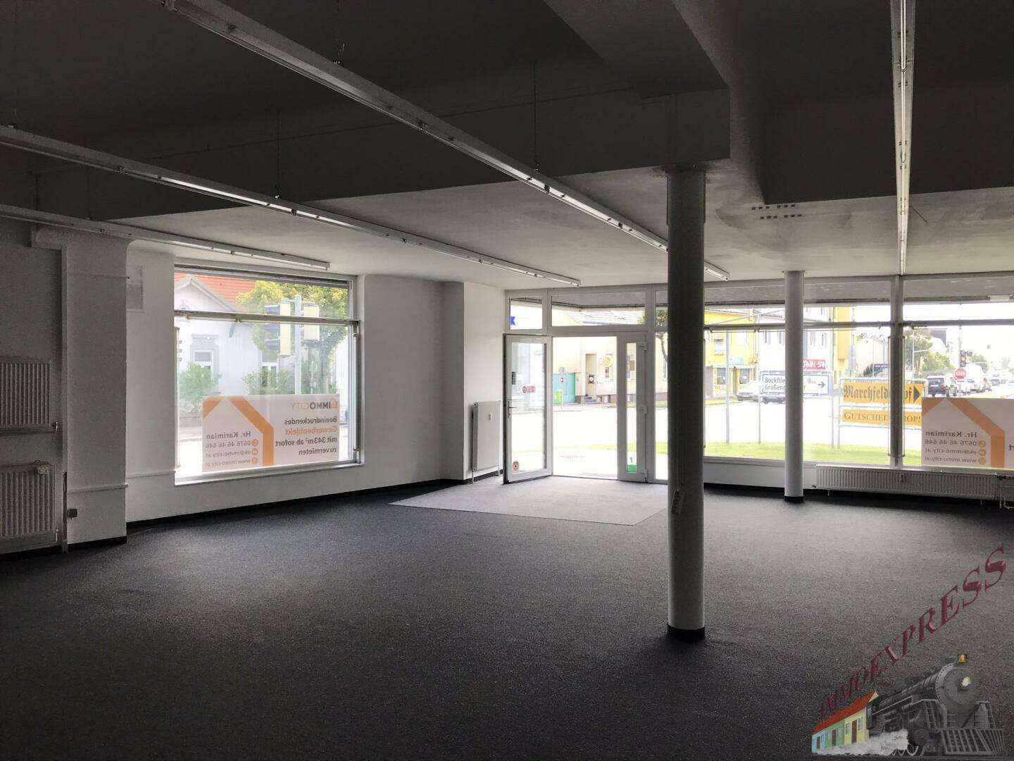 Weitläufiger Ladenbereich mit Teppichboden, Säulen und großen Fenstern zur Straße hin.